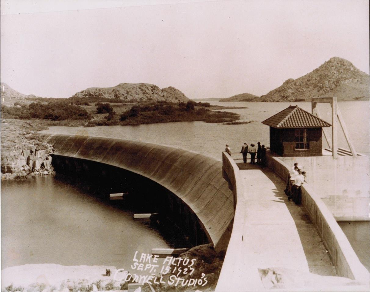 A historical black-and-white photograph of a curved dam with a small control building, surrounded by water and hills in the background, taken in 1925.