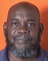 A close-up portrait of a man with a bald head and a full beard, wearing a dark shirt, against an orange background.