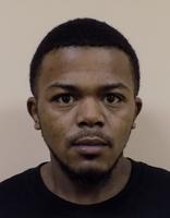 A close-up mugshot of a young man with short black hair and a serious expression, wearing a black shirt.