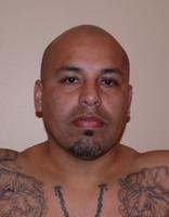 A close-up portrait of a bald man with tattoos on his arms, looking directly at the camera with a neutral expression.