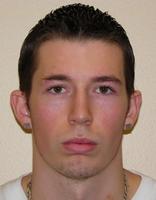A close-up portrait of a young man with short, slicked-back hair and a neutral expression, wearing a white shirt and gold earrings.