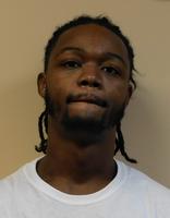 A young man with braided hair is looking slightly downwards, wearing a plain white t-shirt against a neutral background.