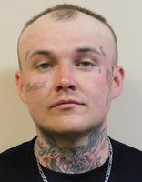 A man with a shaved head and numerous facial tattoos, wearing a black shirt and a silver chain, looking directly at the camera.