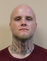 A bald man with tattoos on his neck and a serious expression is facing the camera, wearing a maroon shirt.
