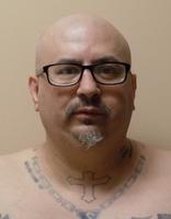 A close-up portrait of a bald man wearing glasses, with visible tattoos on his chest and neck, against a plain background.