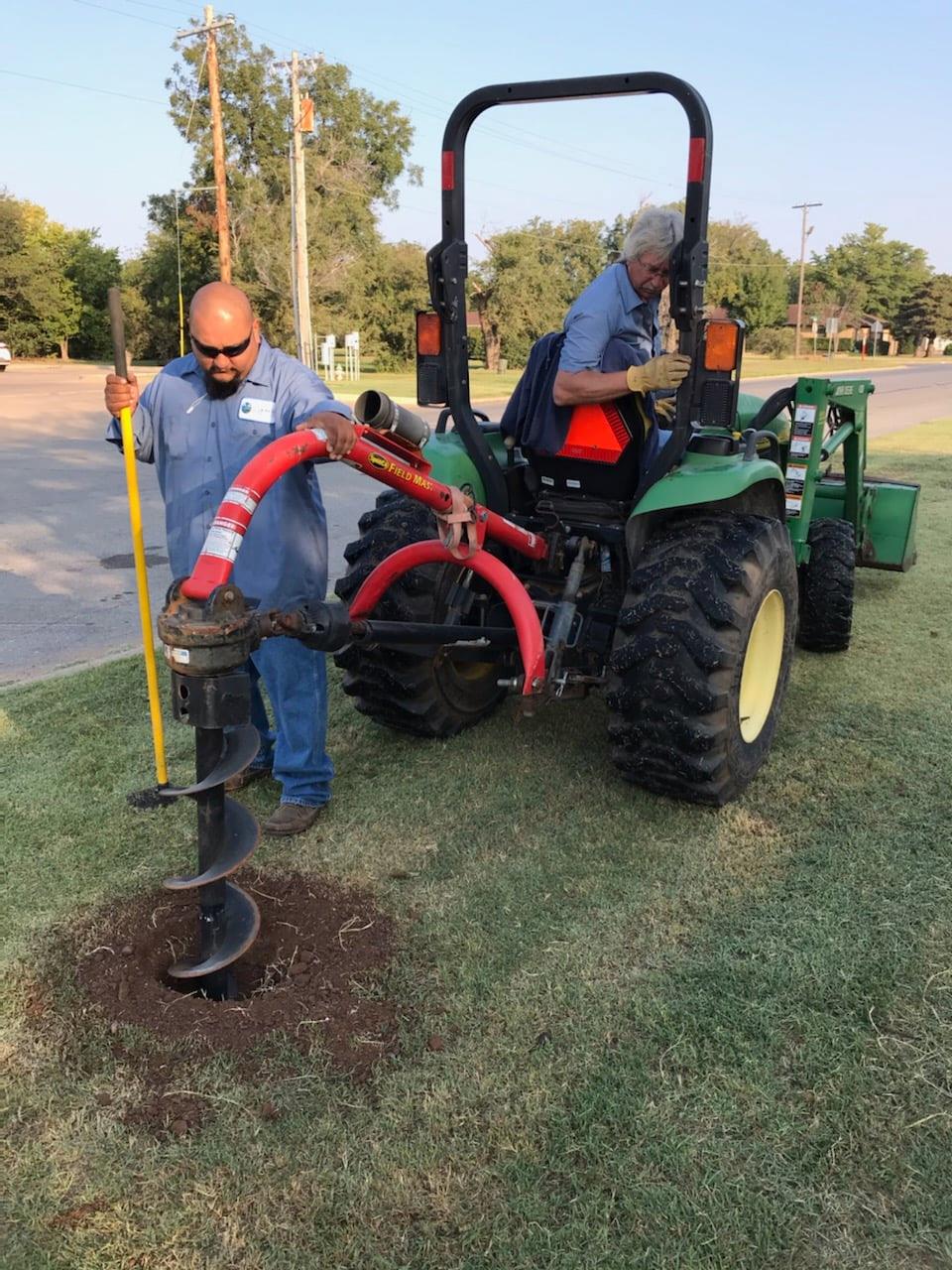 Two workers operating a tractor-mounted auger to drill a hole in a grassy area. One worker is standing and guiding the auger, while the other is seated on the tractor, overseeing the equipment.