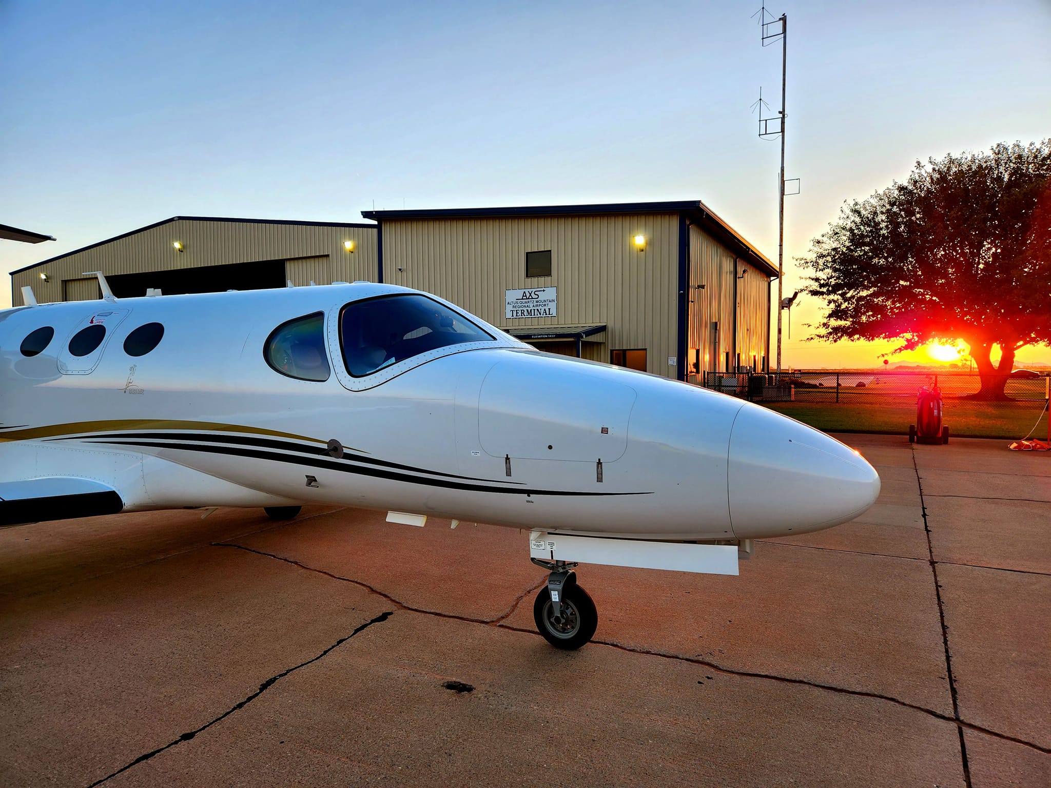 Small white aircraft parked on the tarmac outside the Altus Municipal Airport terminal, with the sun setting behind the building and casting a warm glow over the scene.