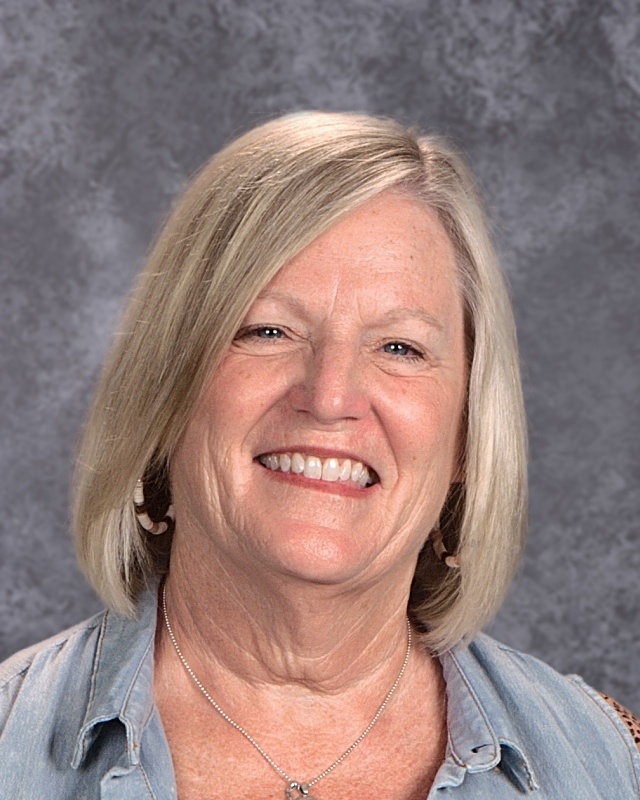 A woman with blonde hair wearing a blue shirt, smiling and standing against a neutral background.