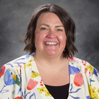 Woman with short hair smiles at camera; wears floral-patterned top over black shirt against gray background.