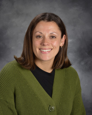 A woman with short brown hair, smiling, wears a green cardigan over a black top against a gray background.