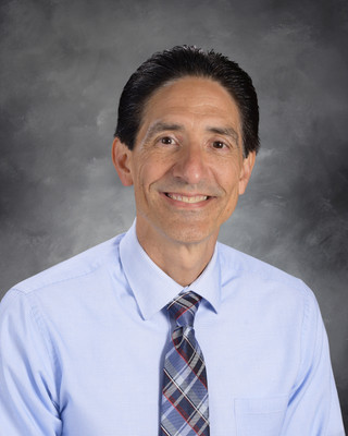 A man smiles, standing against a gray background. He wears a light blue shirt and a plaid tie.