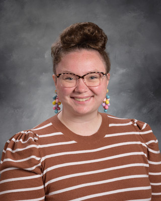 Woman with glasses and a bun hairstyle, smiling, wearing a brown and white striped shirt. Background is a solid gray.