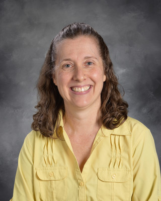 A woman with brown curly hair wearing a yellow shirt poses for a photo against a gray background.