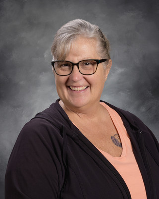 A woman with gray hair wearing glasses, a black top, and a pink shirt underneath, smiling against a gray background.