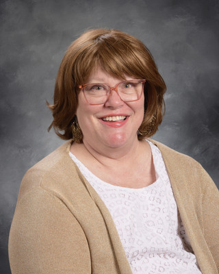 A woman with short brown hair, wearing glasses and a beige cardigan over a white blouse, smiles for the camera against a gray background.