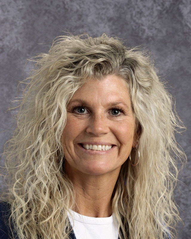 A smiling woman with long, curly blonde hair, wearing a dark blazer over a white shirt, poses for a professional headshot against a gray background.