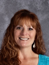 A smiling woman with long, curly red hair, wearing a turquoise top and earrings, set against a gray background.