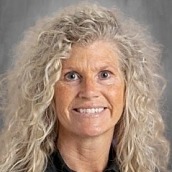 A woman with blonde, curly hair in a black shirt, standing in front of a gray background, smiling.