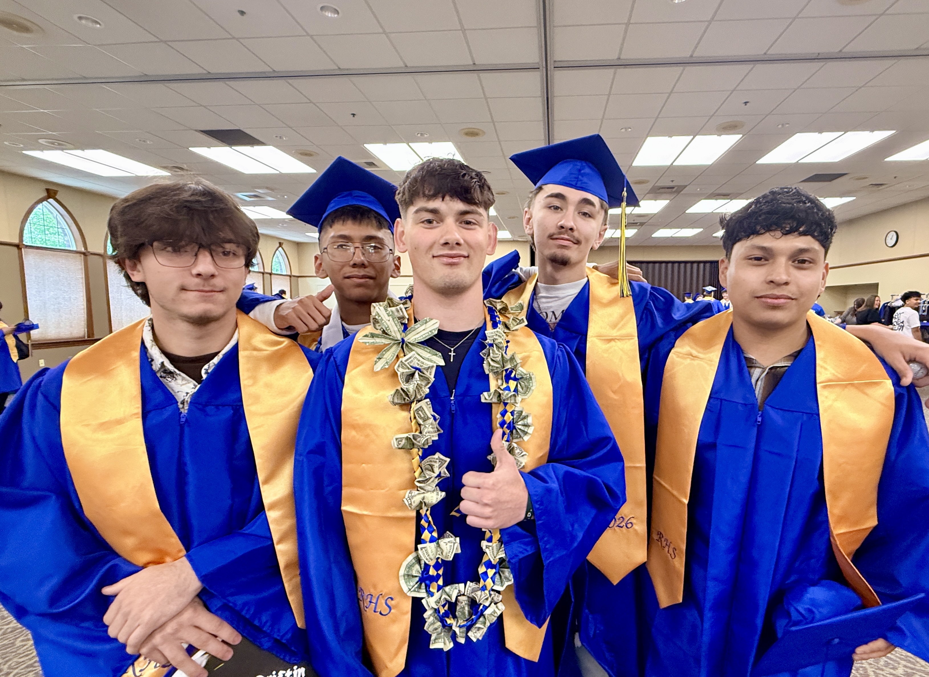 Five individuals in blue and gold gowns and caps stand in a room. One person poses with a thumbs-up.