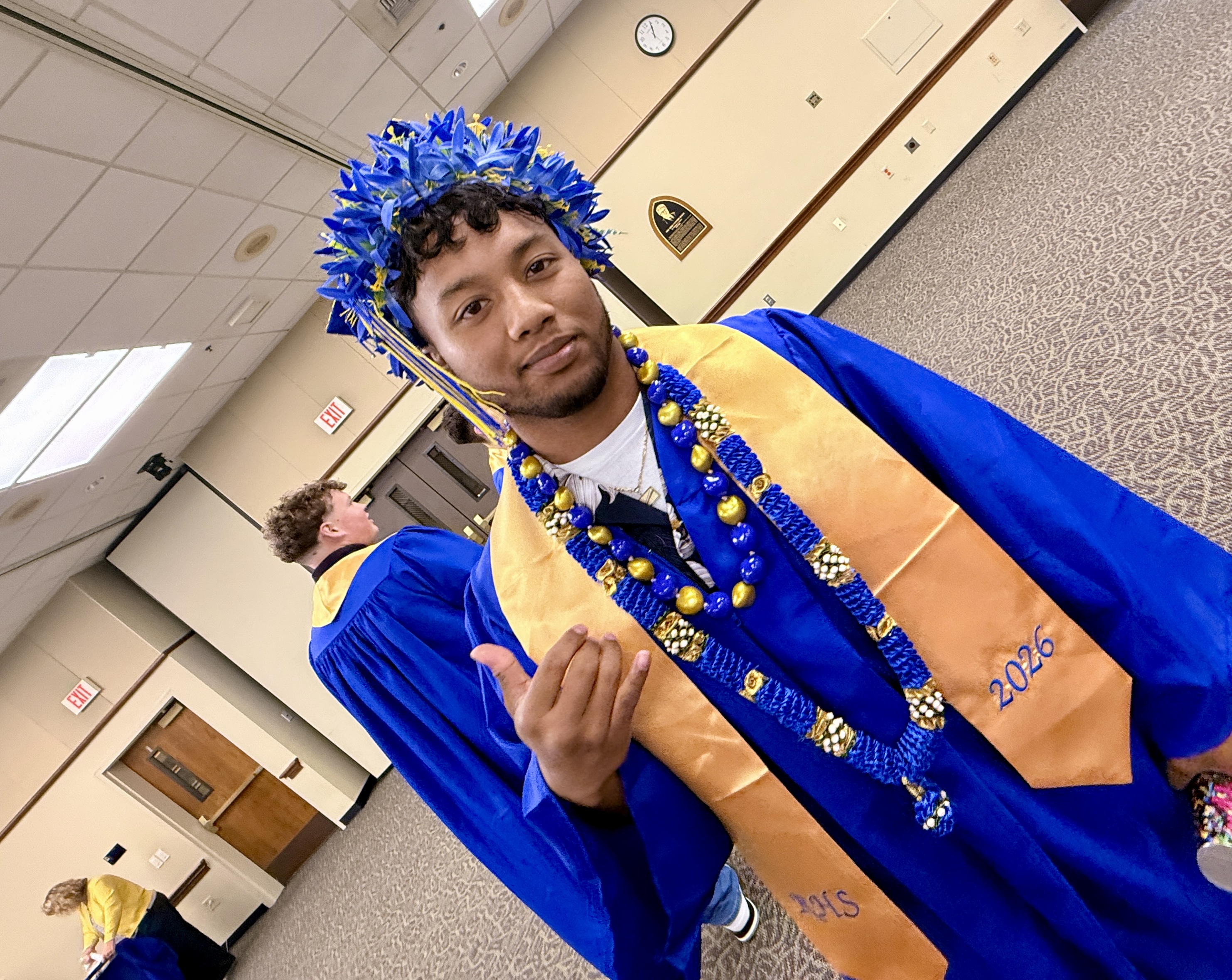 A man in a blue and gold graduation gown, wearing a blue graduation cap, poses for a photo.