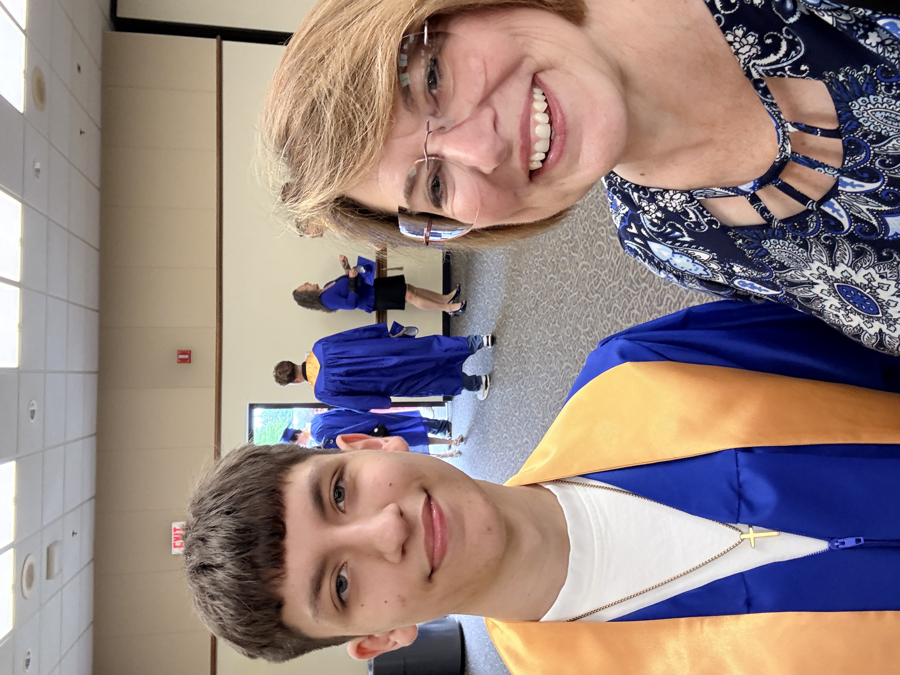 A young man and an older woman stand together, smiling, with a school hallway in the background.
