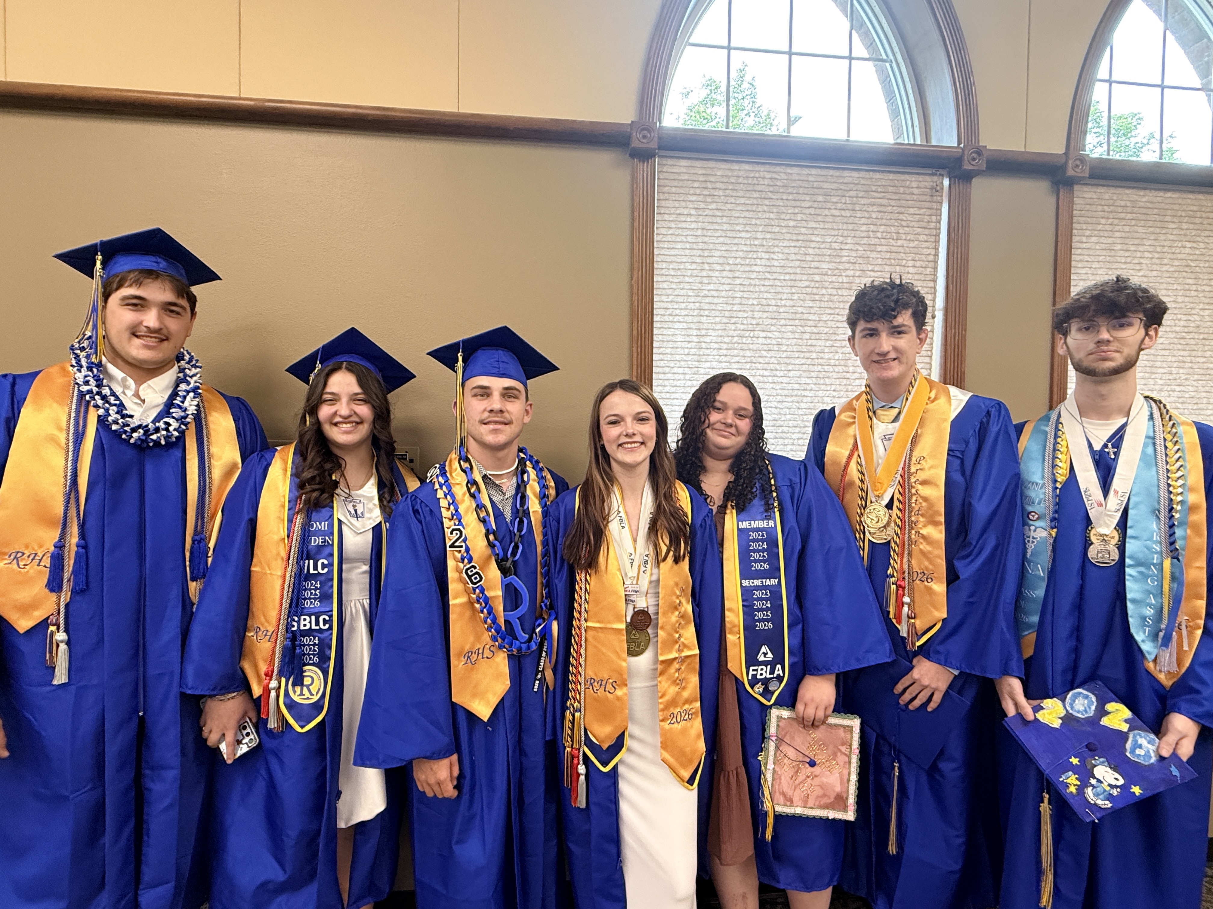 Graduates in blue caps and gowns stand together, some holding diplomas, in front of arched windows.