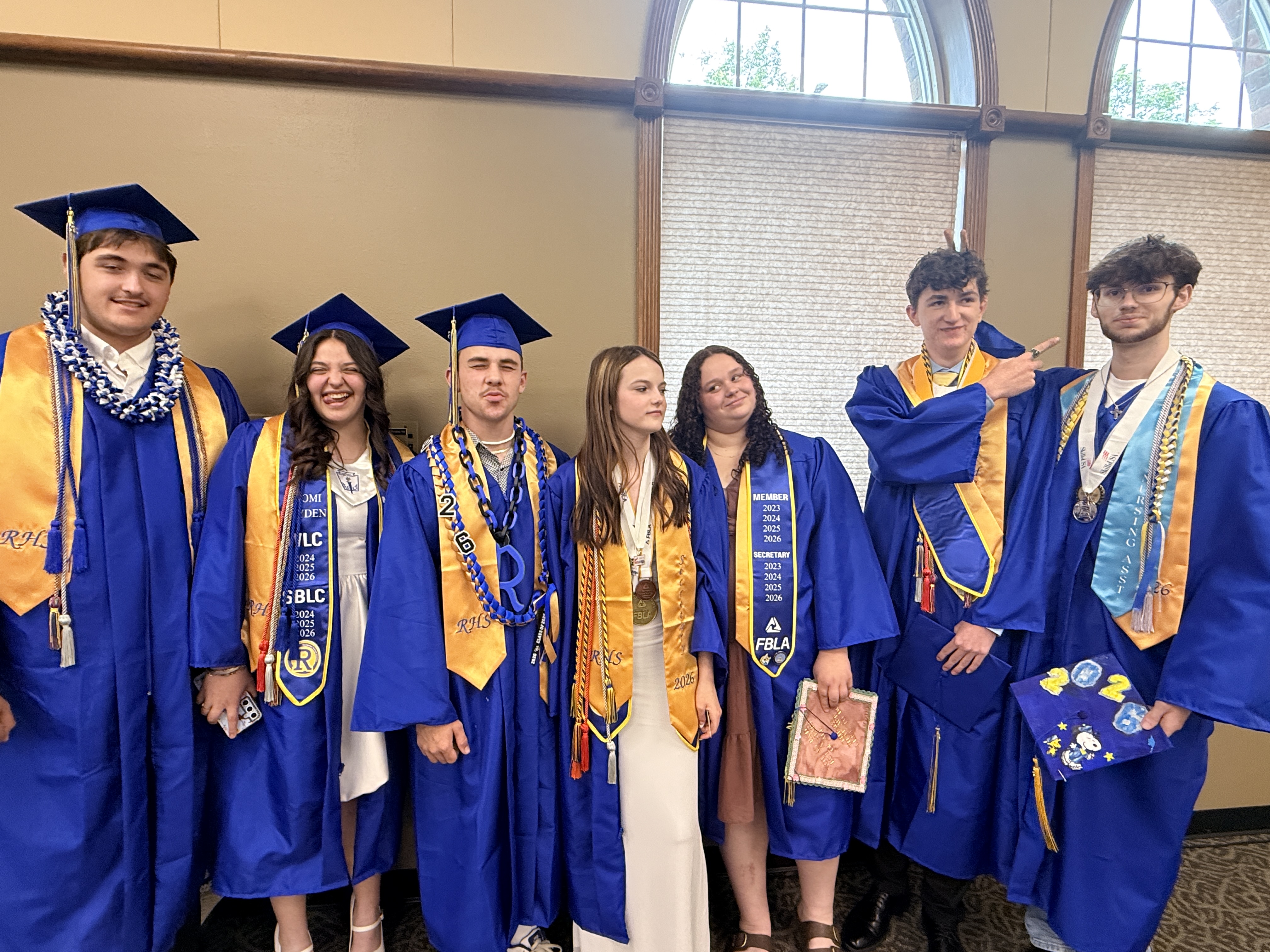Several students in blue caps and gowns stand together, smiling. Some hold books and certificates. One wears a white dress. Behind them, an arched window lets in natural light.