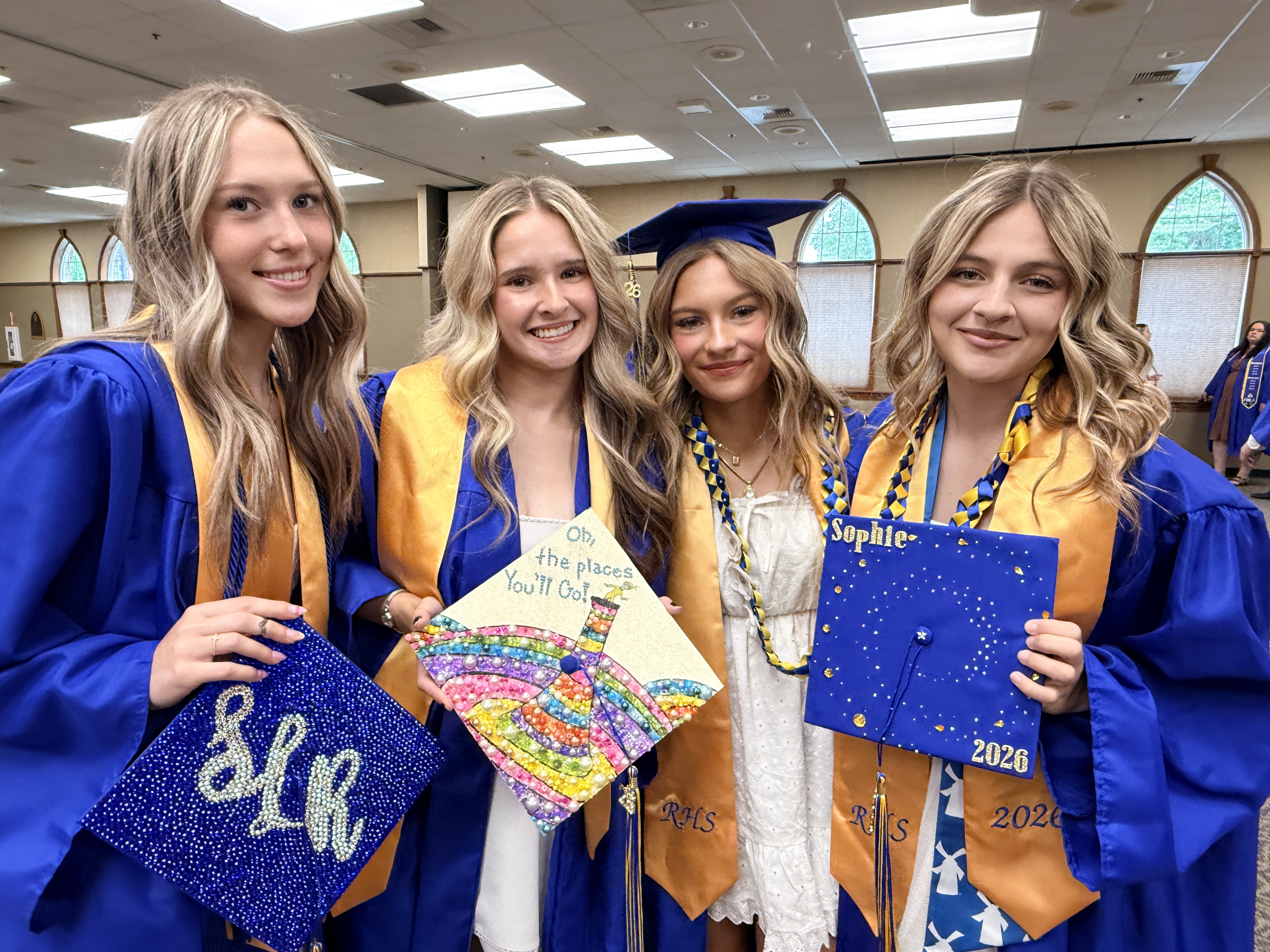 Four women in blue and gold graduation robes. Two hold graduation caps. One holds a colorful paper craft.