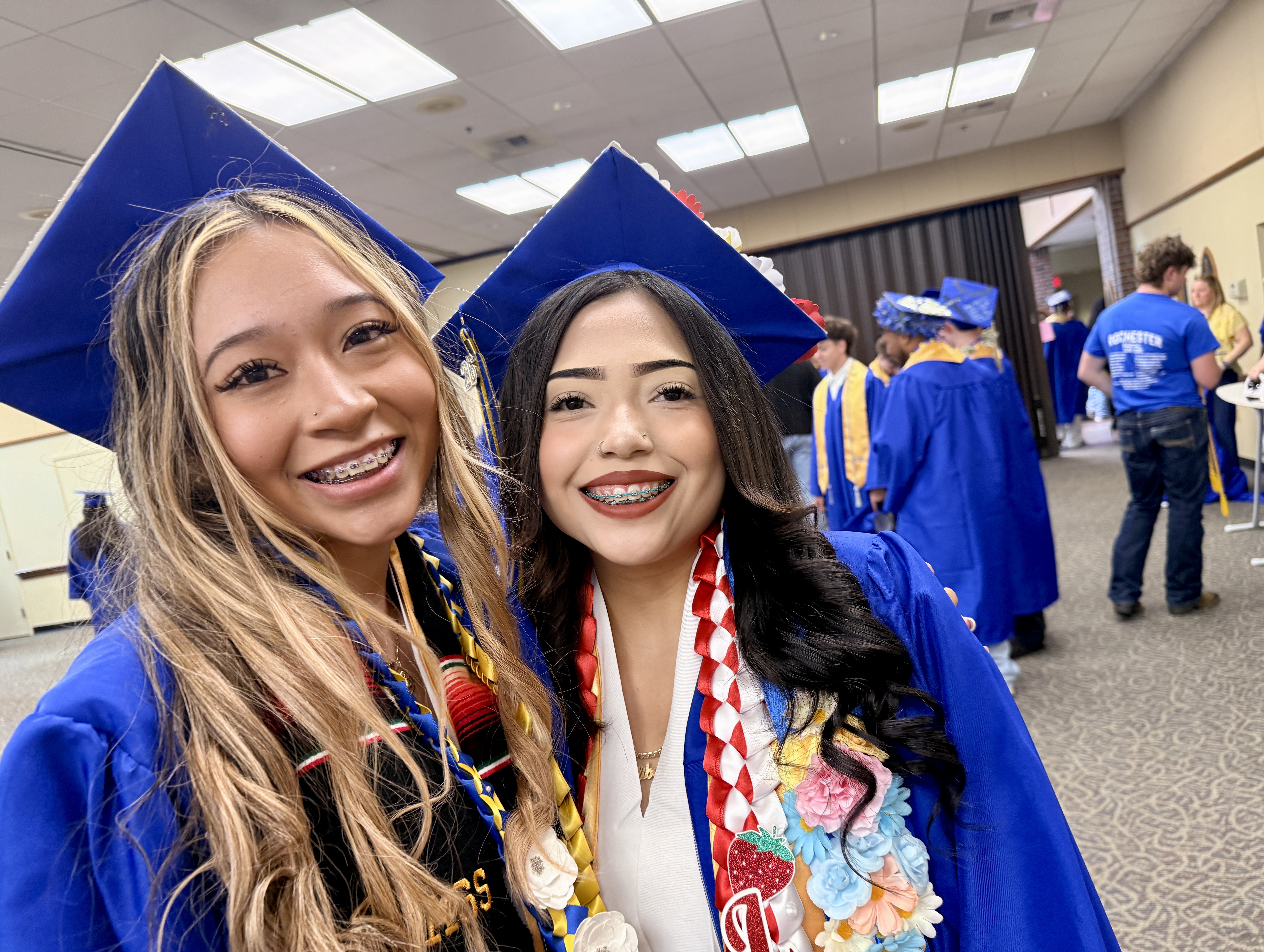 Two women in blue graduation gowns and caps smile in a hallway, with other students in blue gowns behind them.