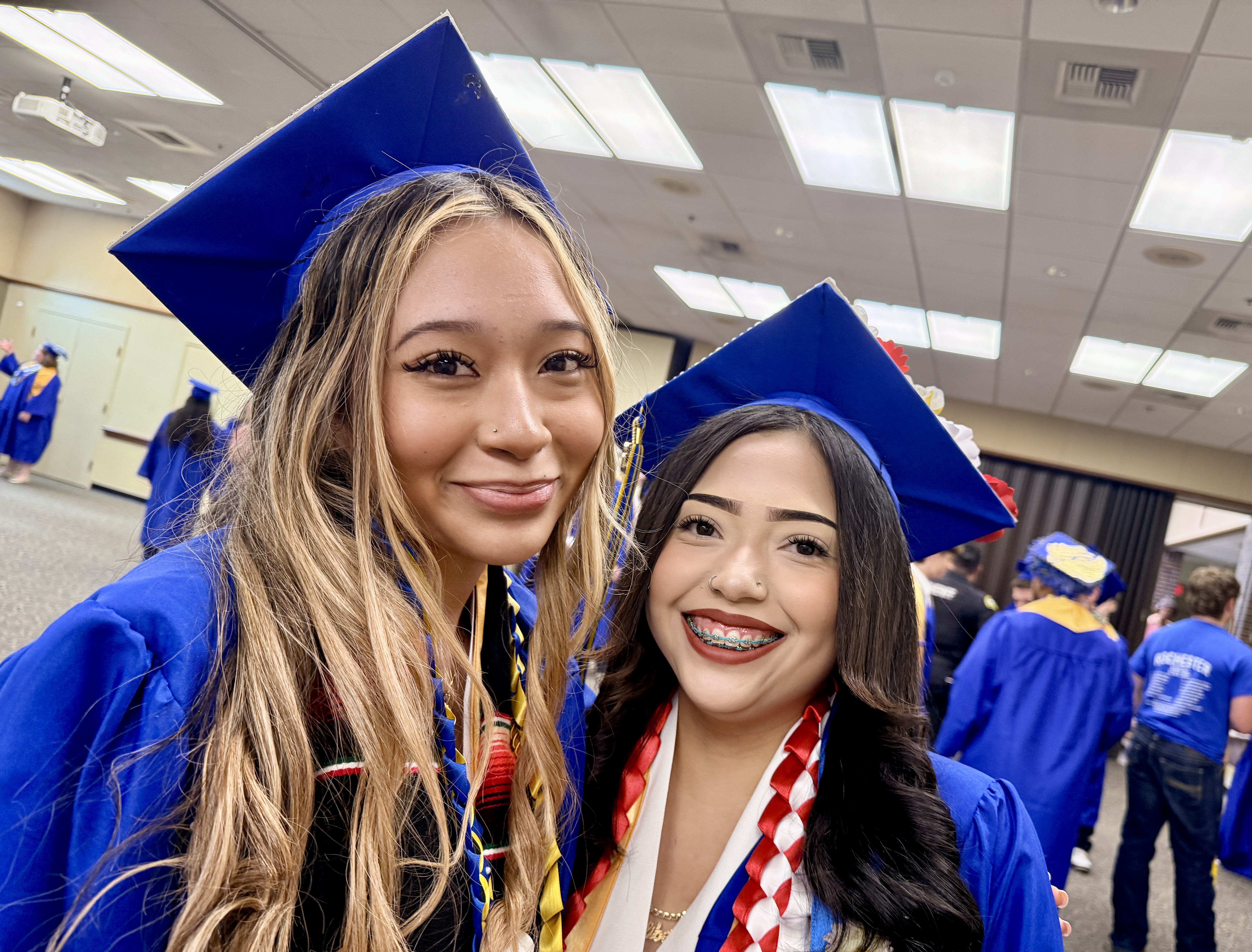 Two women in blue graduation gowns and caps pose for a photo. They smile and have long hair.