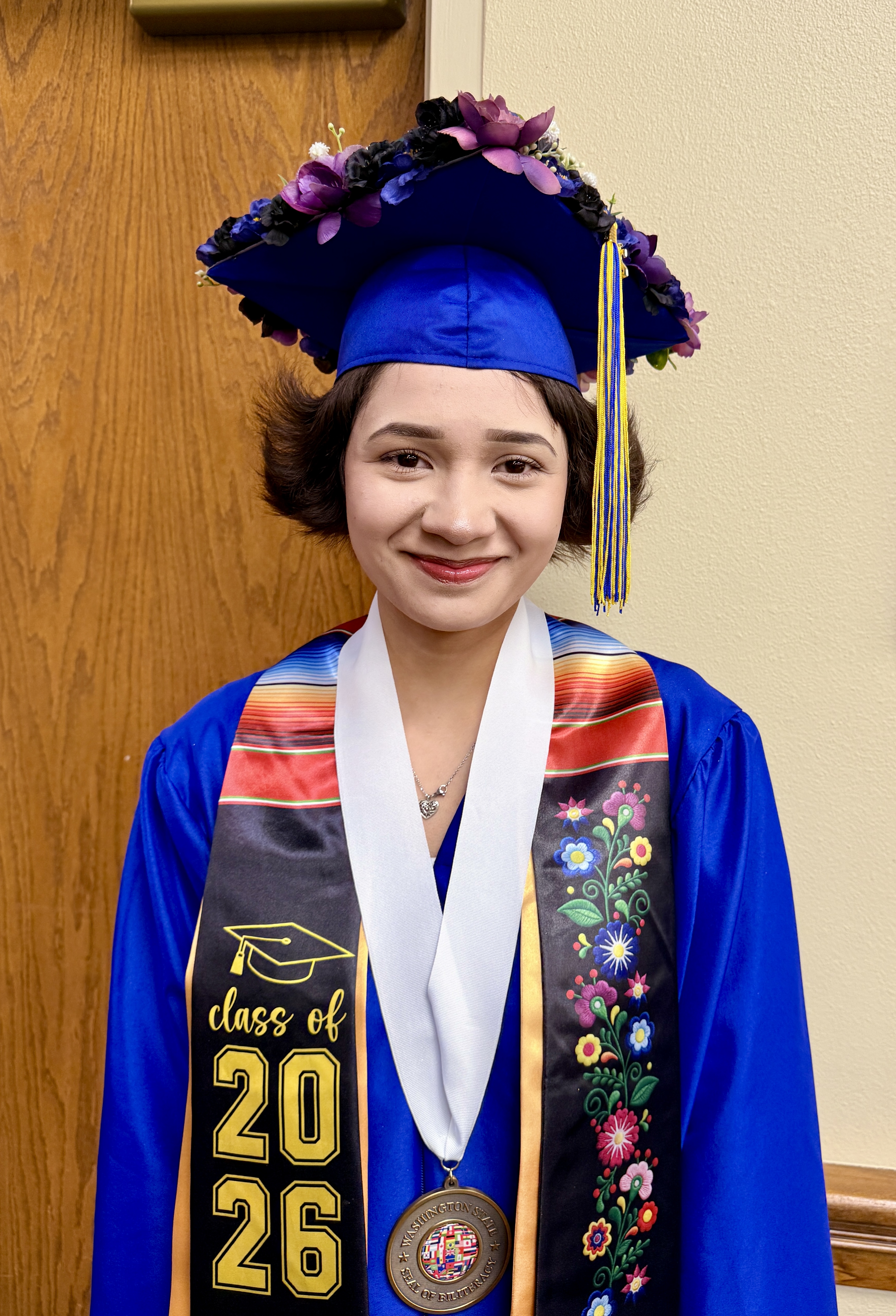 A smiling woman in a blue gown and cap, with a sash reading "Class of 2026", stands by a wooden door.