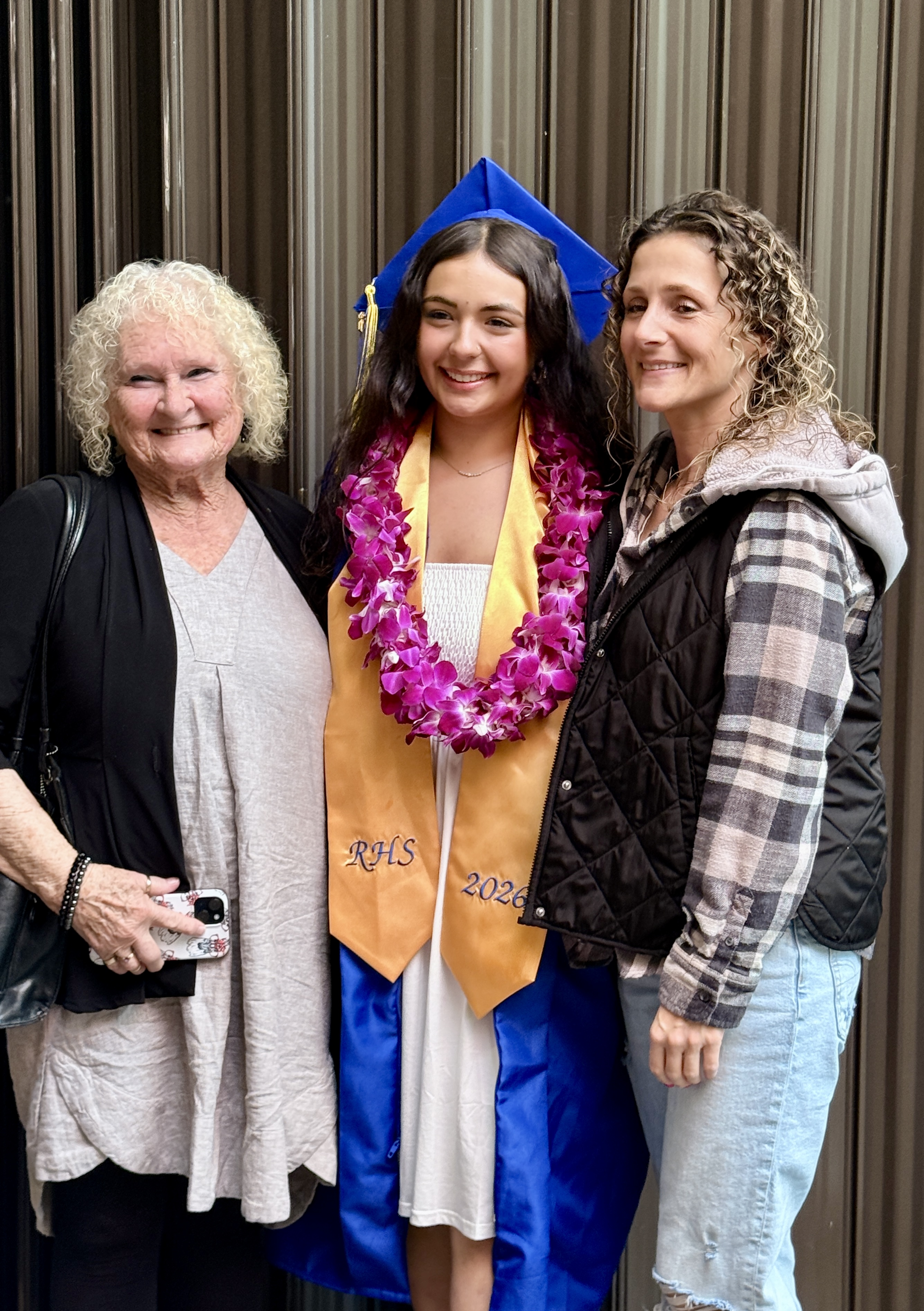 Three people smile, including a woman in a graduation gown with a lei, a woman in a jacket, and a woman in a gray top.