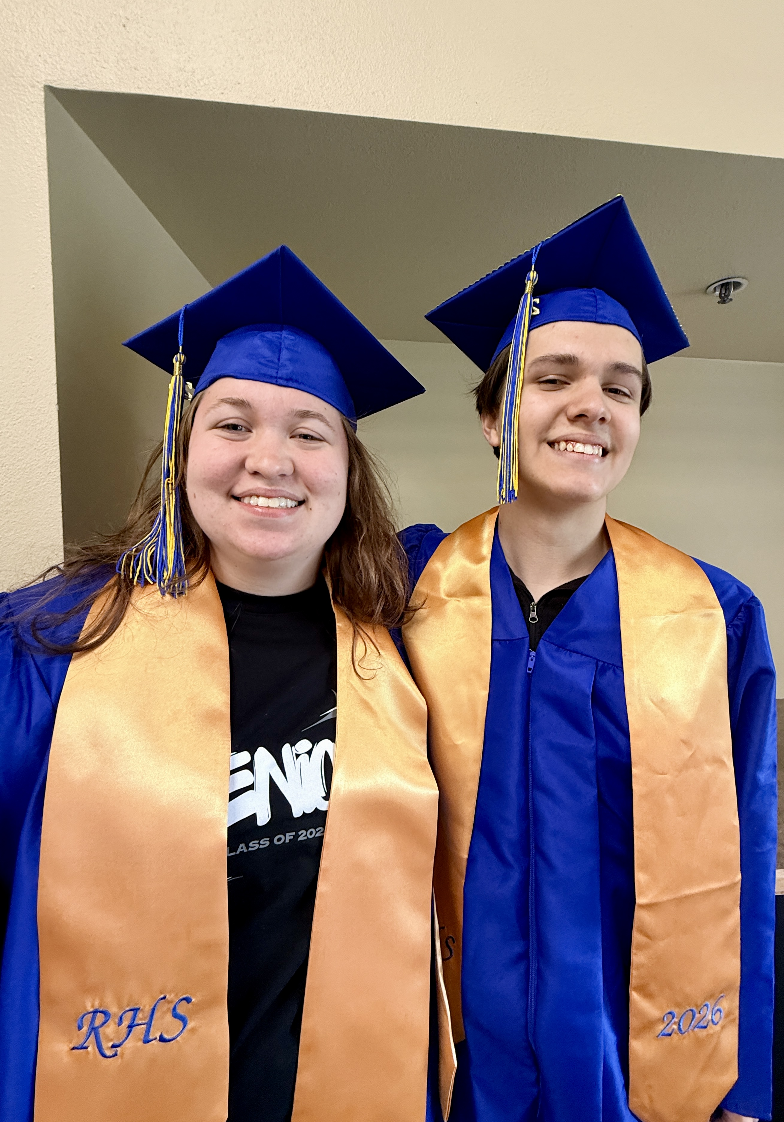 Two people in blue graduation gowns and blue hats. The woman's gown has an orange sash.