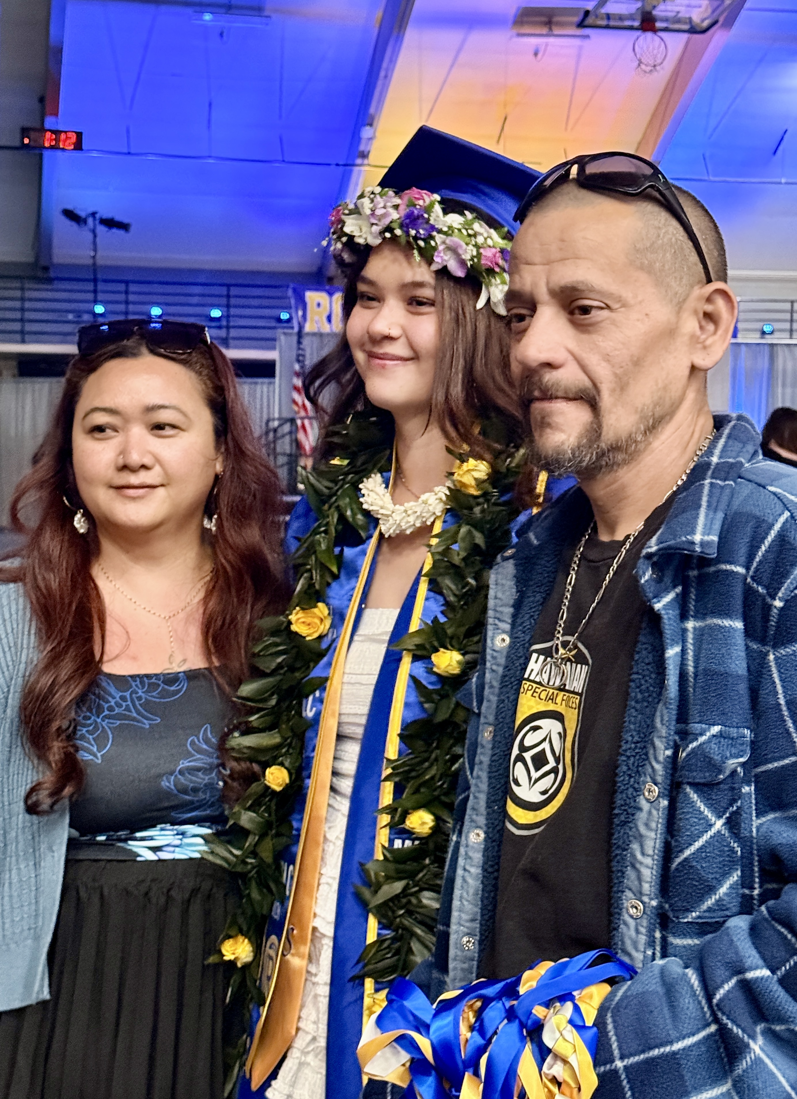 Two women and a man stand close together in a room. The man wears a plaid jacket. A woman wears a blue gown and lei.