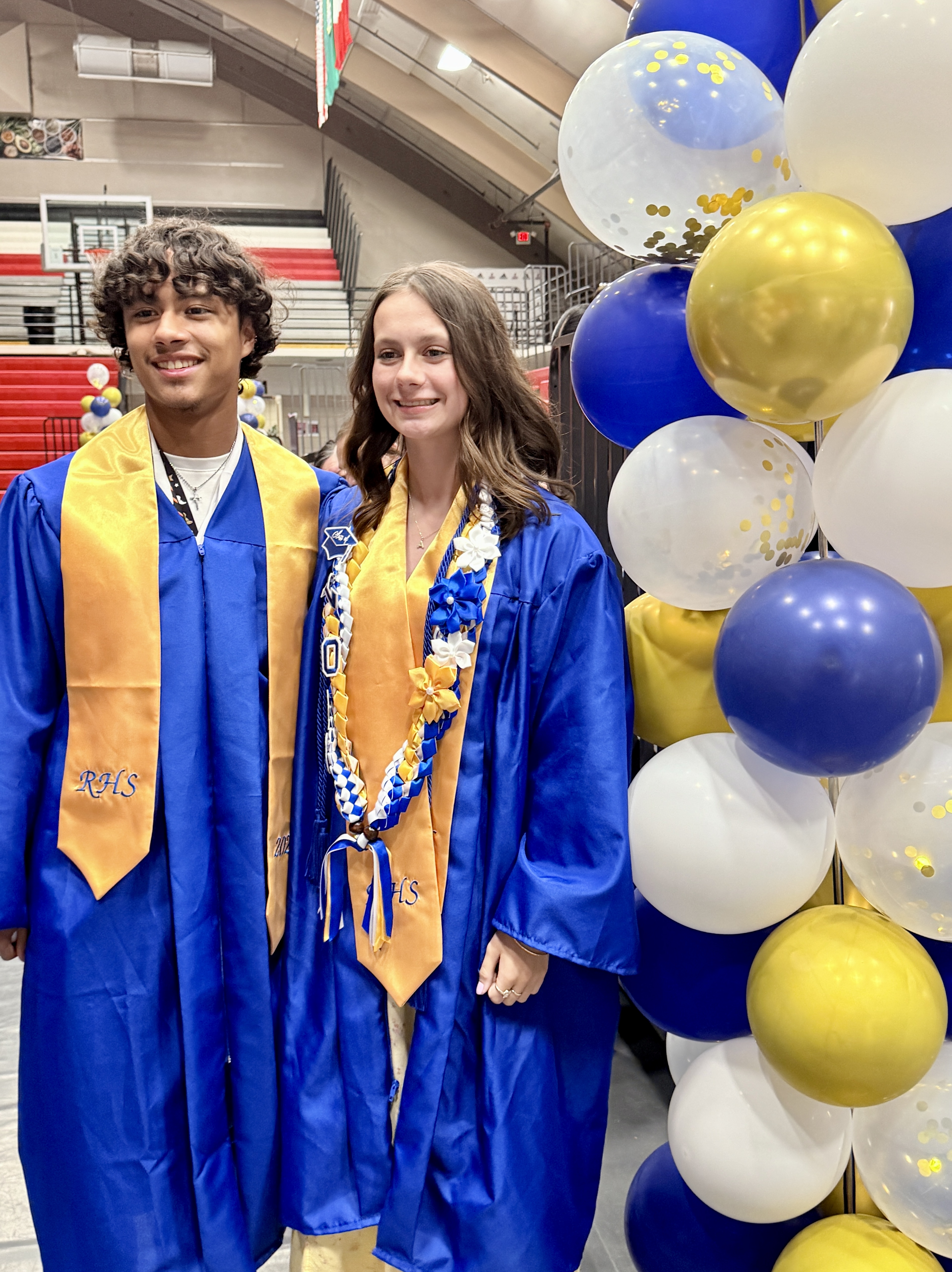 Two individuals in blue gowns, one with a gold sash, smile next to a blue and white balloon arch.