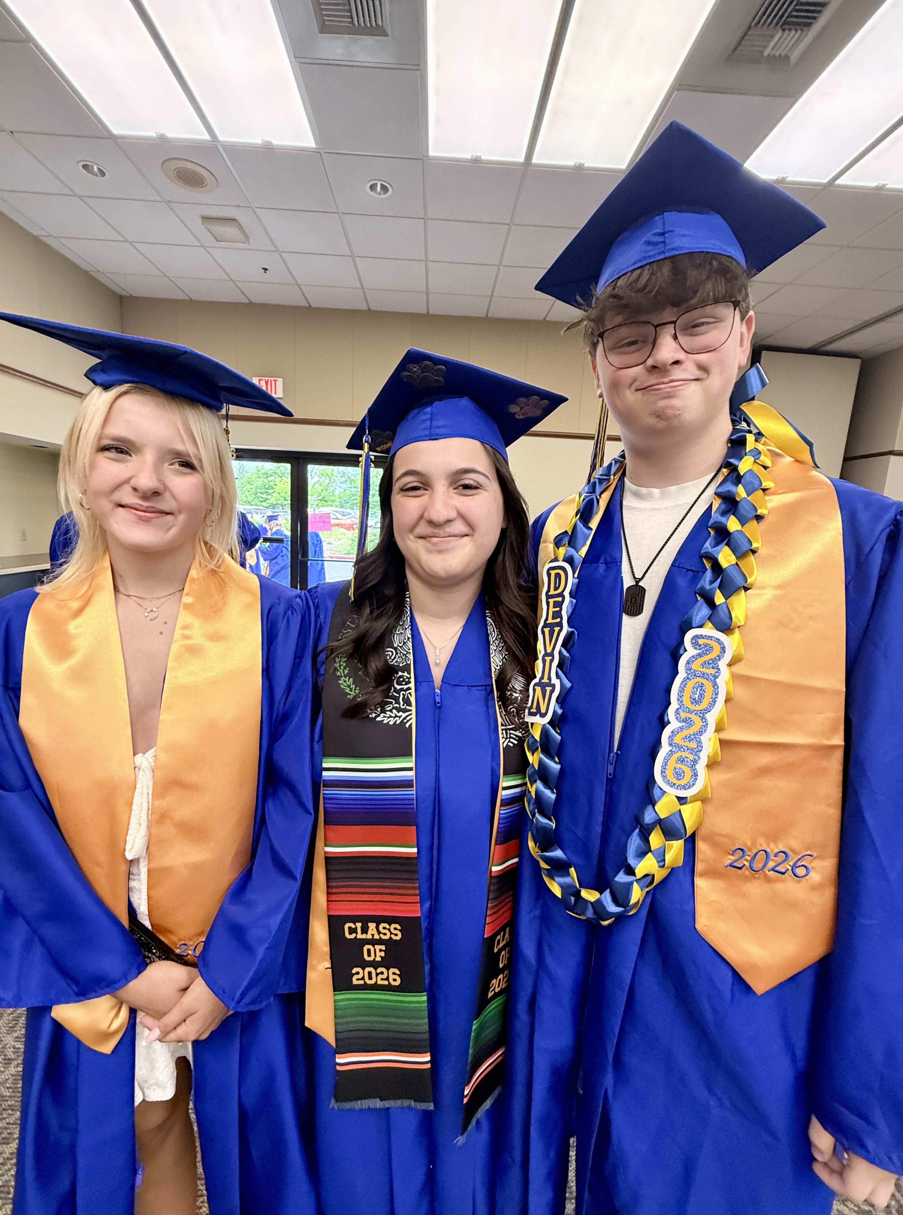Three people in blue graduation gowns and hats, with one wearing a sash that says 2026.