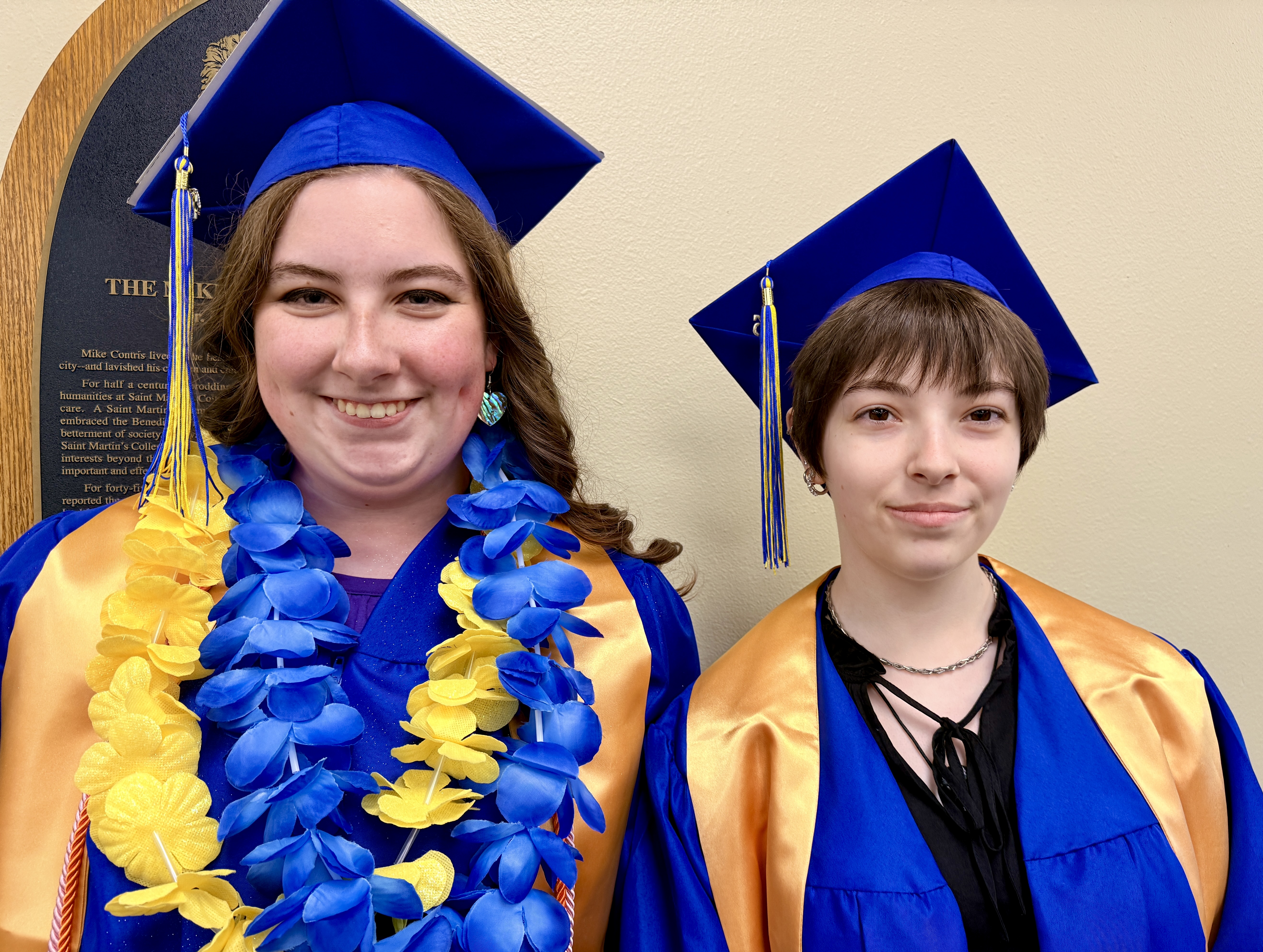 Two individuals in blue and gold graduation gowns. One has a blue and yellow garland.
