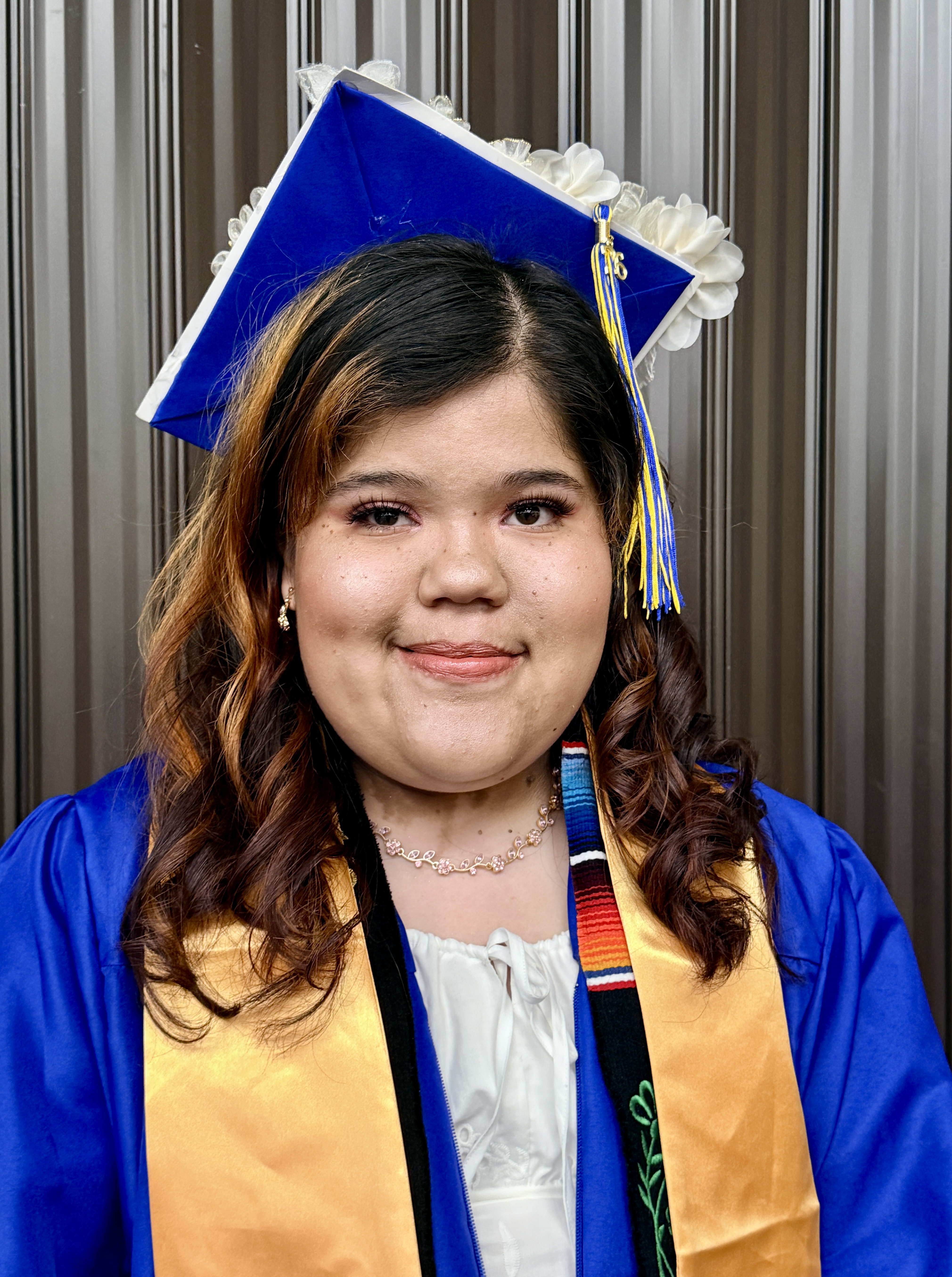 A woman in a blue gown and cap, smiling, stands with a gray background. She wears a necklace and sash.