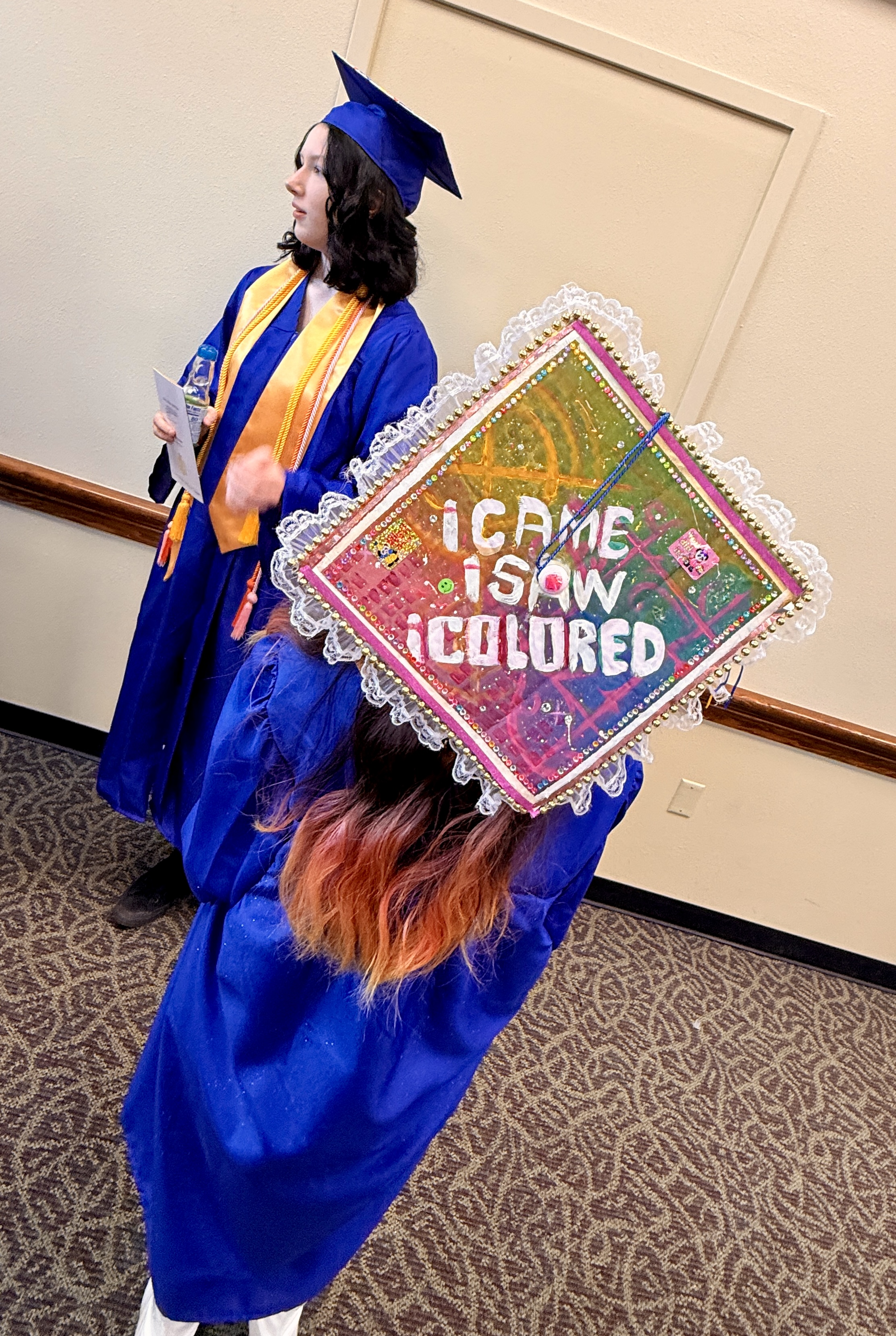 Two women in blue graduation gowns, one with a cap reading "I Came I Saw I Coloured." They stand in a corridor.