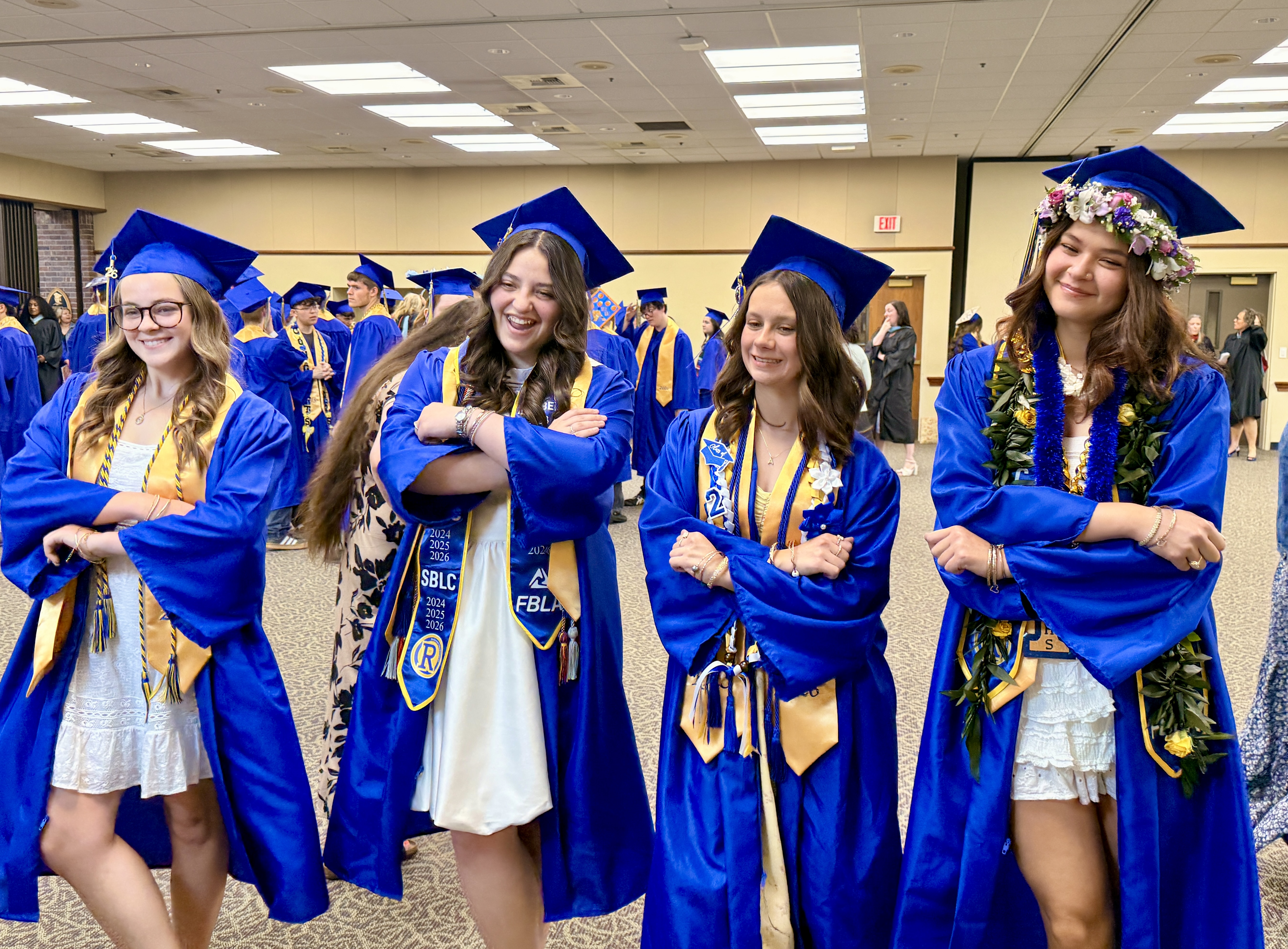 Four women in blue graduation gowns pose for a picture in a room with many other graduates.