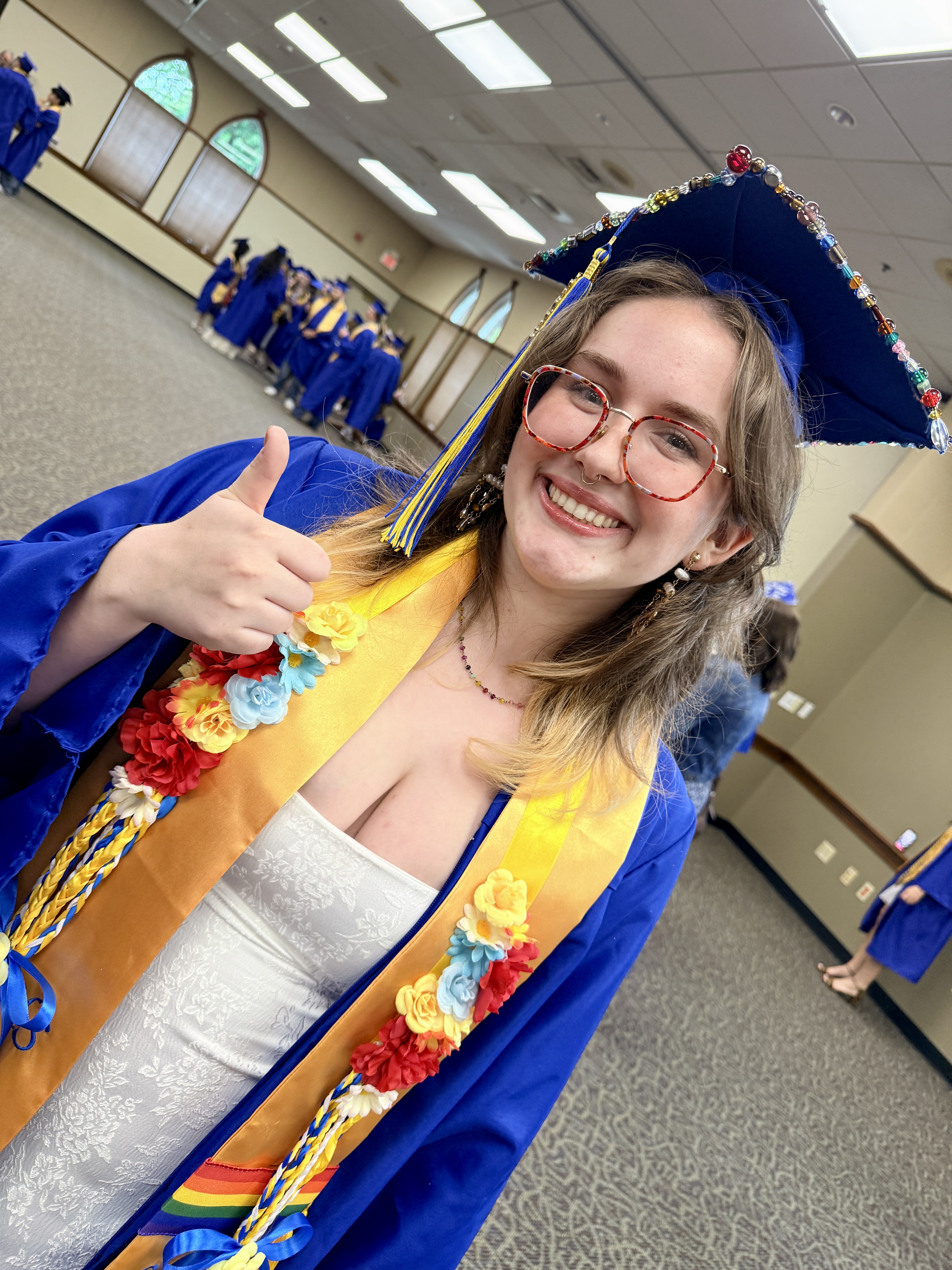 A woman in a blue gown, cap, and glasses gives a thumbs-up. She wears a necklace and a colorful sash.