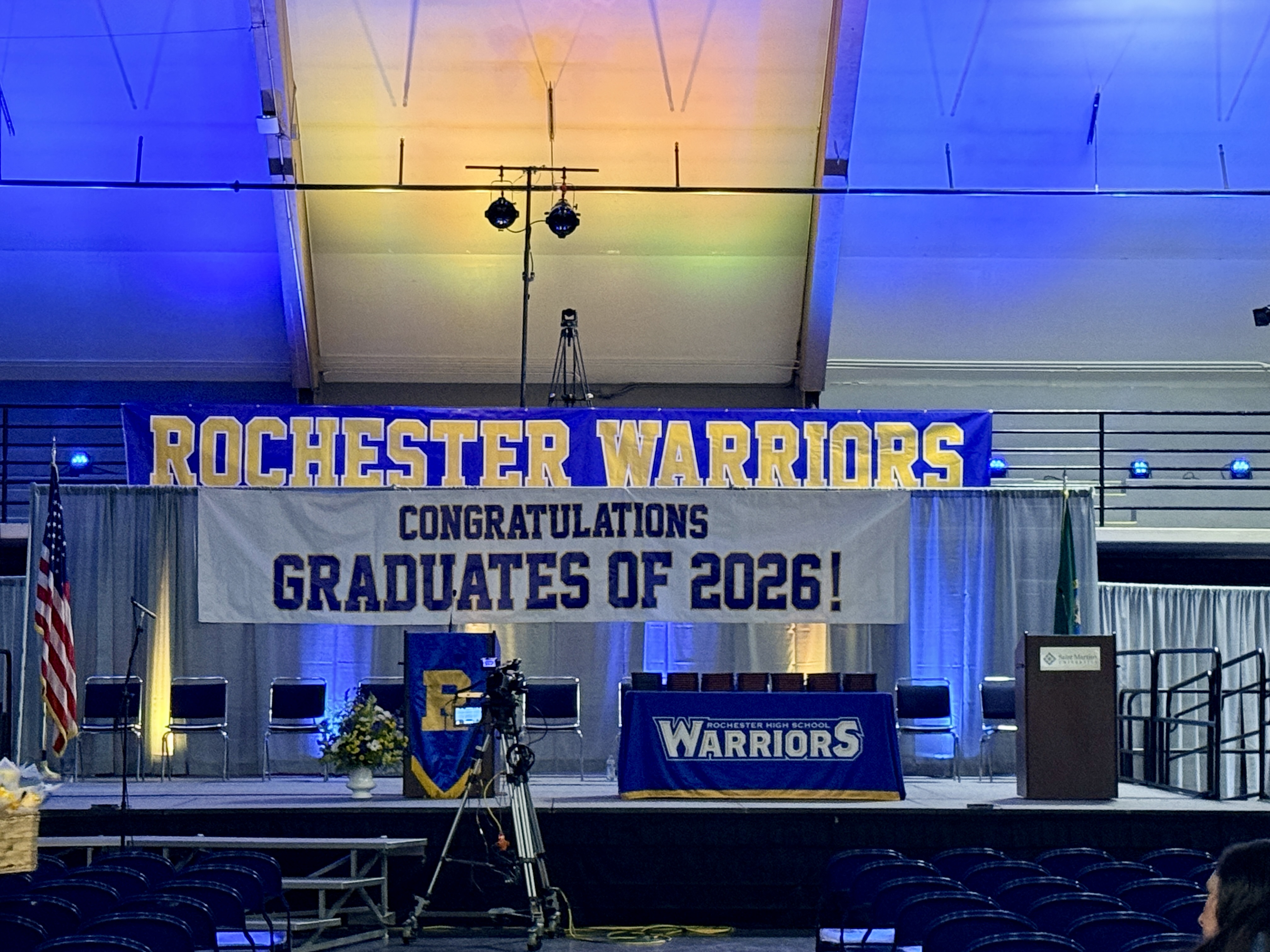 A graduation stage with a large banner reading "Rochester Warriors" and "Congratulations, Graduates of 2026!". A podium is in the center with an American flag.