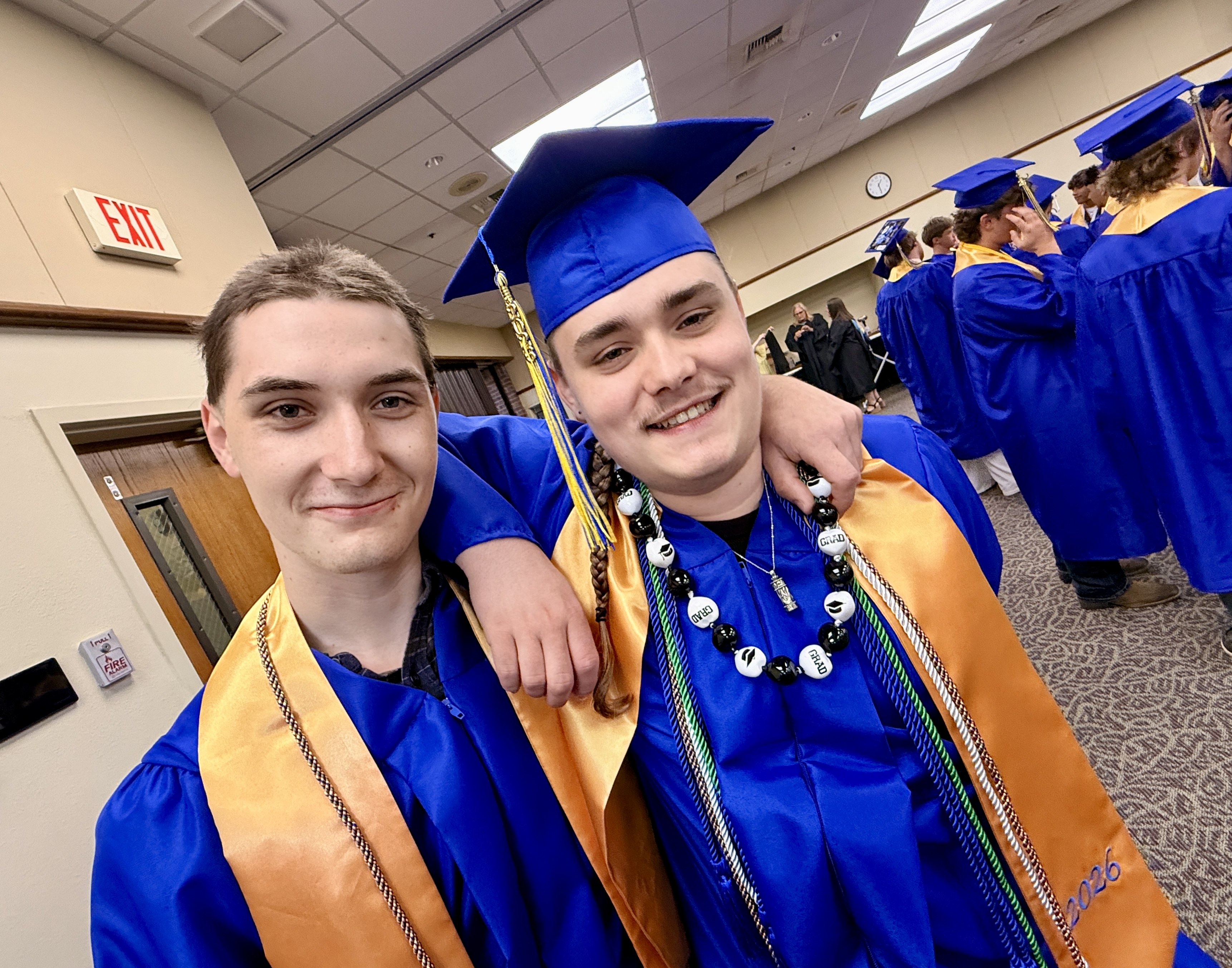 Two men in graduation gowns and caps pose for a picture; one has a necklace and the other has a cap tassel.