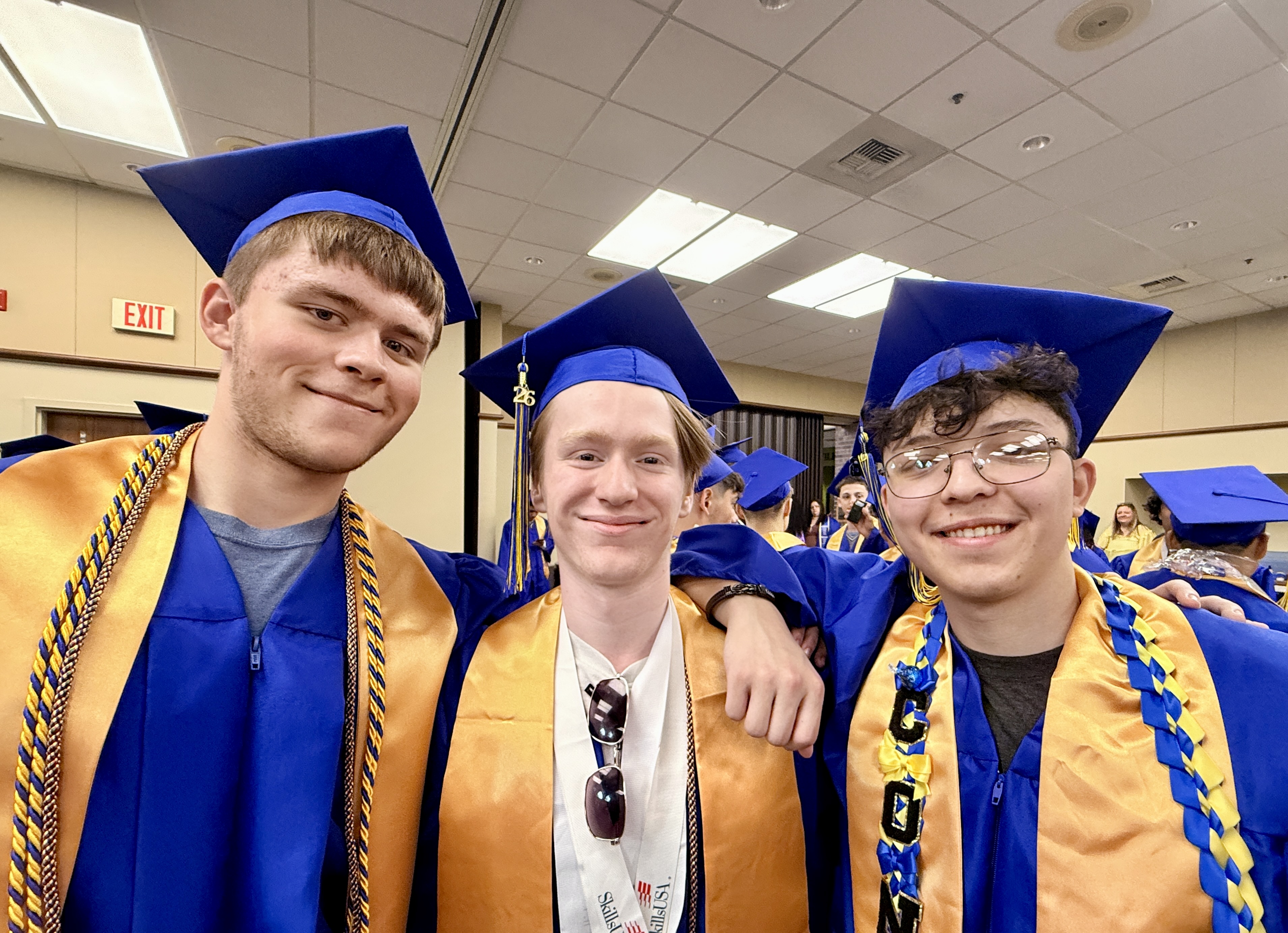 Three young men in blue and gold graduation gowns and caps pose for a photo at an event.