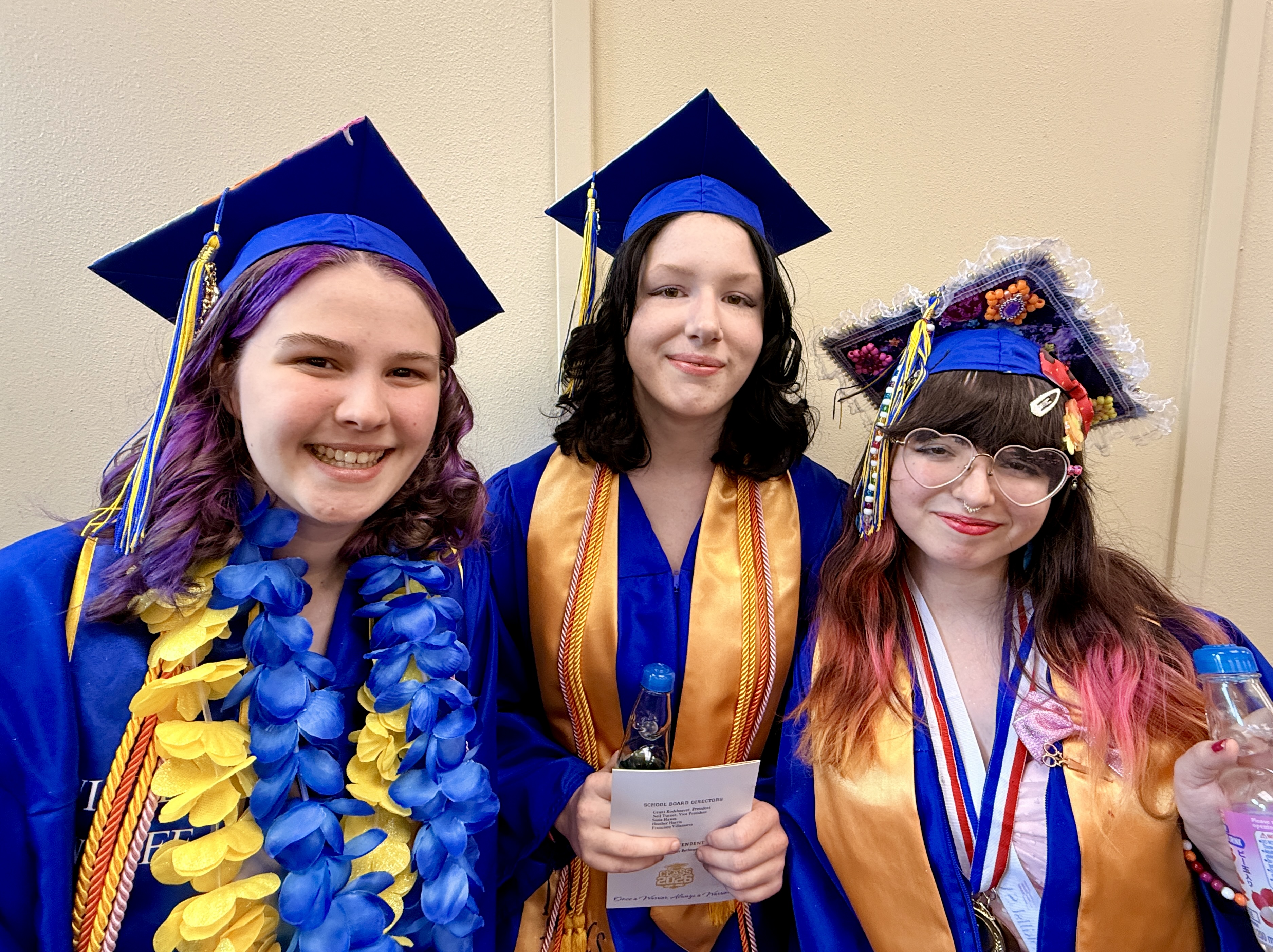 Three women in blue and gold graduation gowns pose together; one has a blue lei and purple hair, another holds a certificate.