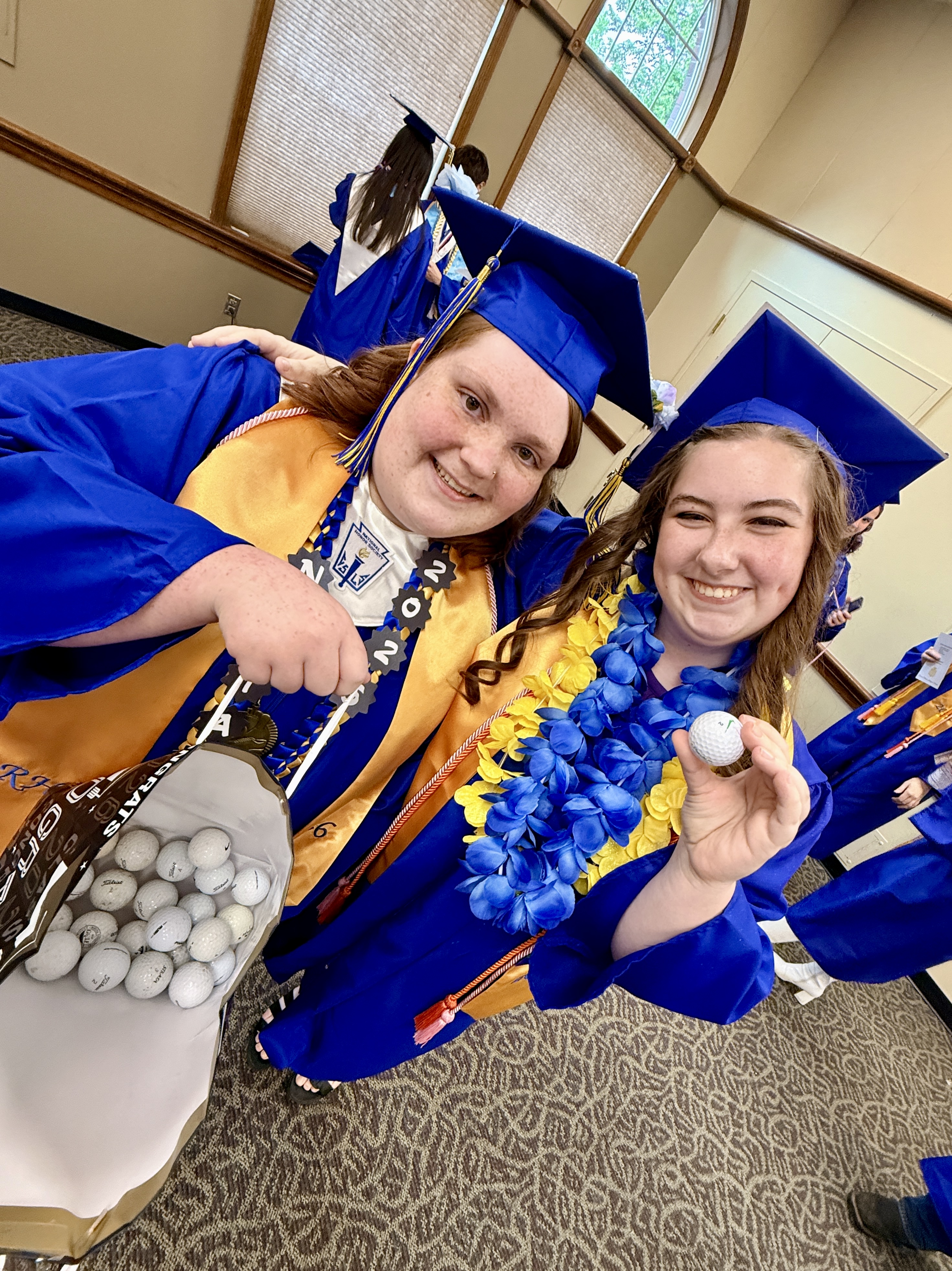 Two women in blue gowns and hats hold a golf bag and ball, posing for a photo.