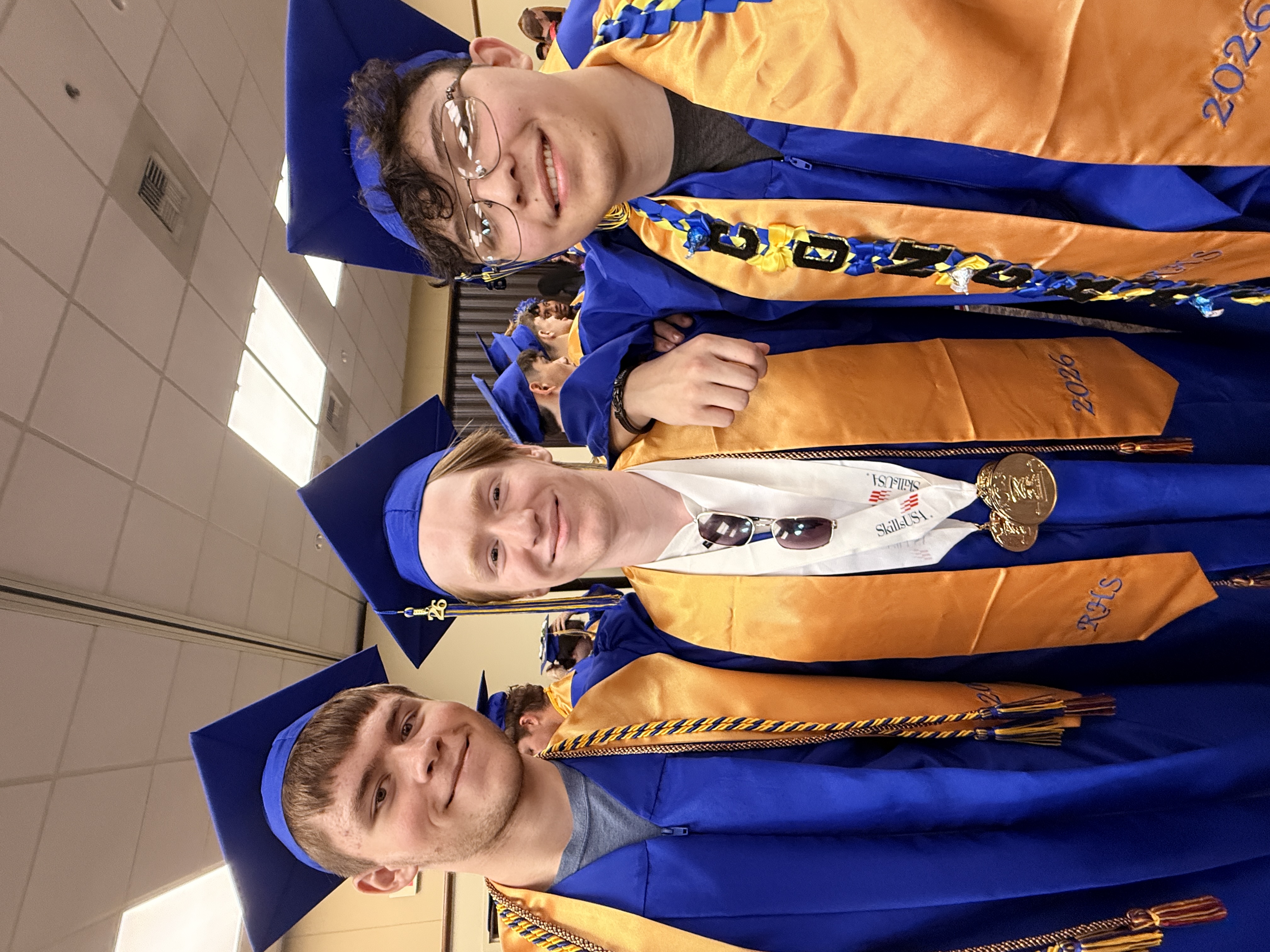 Three young men in blue and gold graduation gowns pose for a photo. The middle man wears a sash with medals.