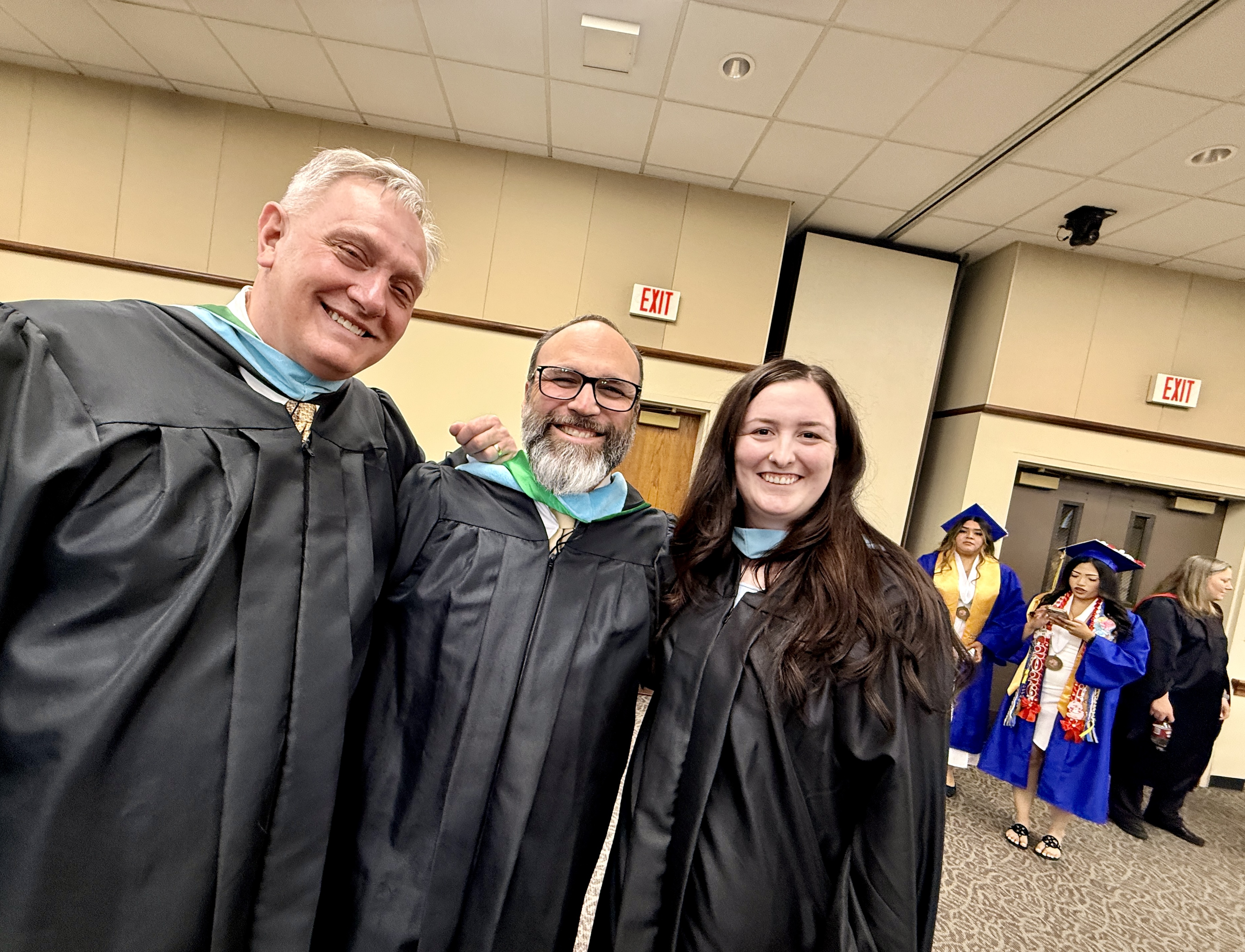 Three people in academic regalia, two men and a woman, standing close together with others in gowns behind them.