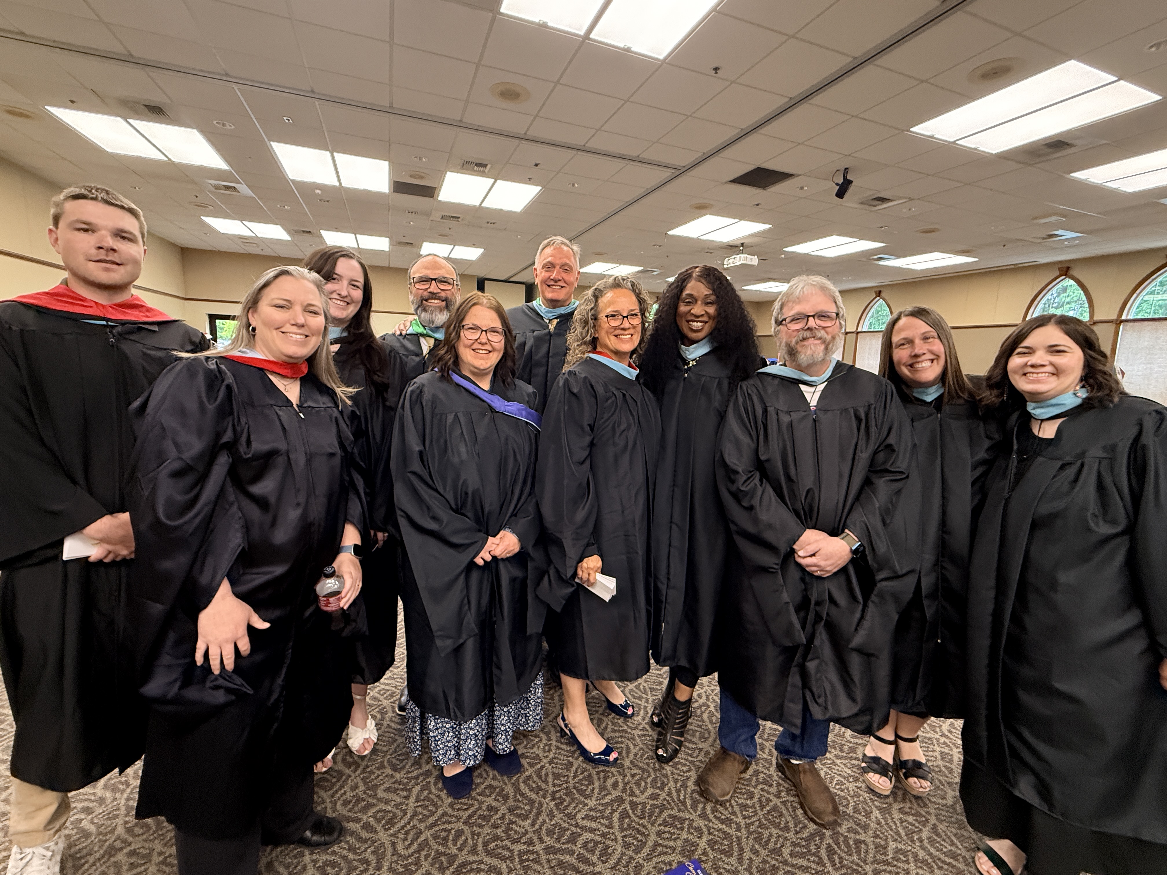 A group of 12 people in black graduation gowns, some in caps, smile for a photo in a room.