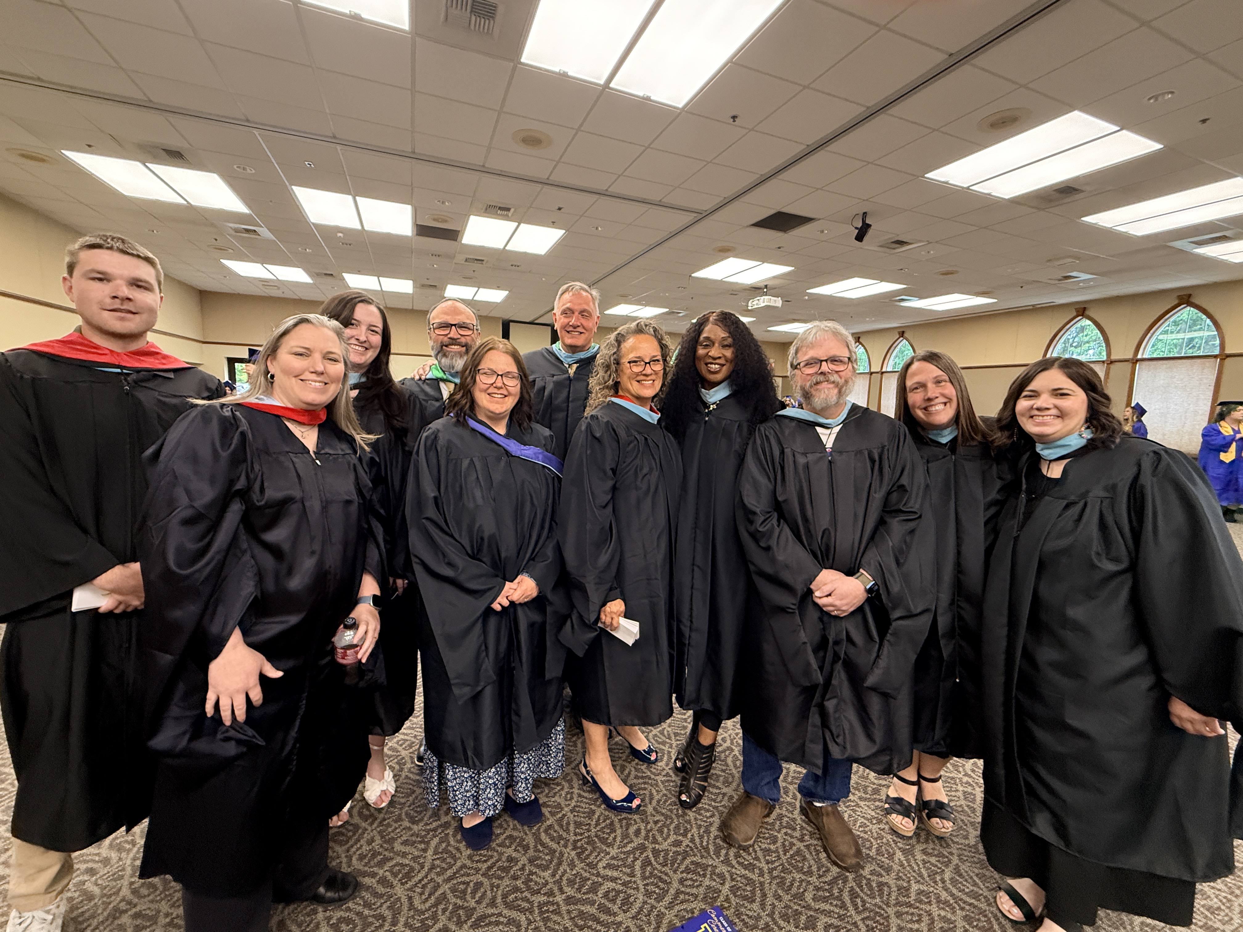 A group of people in black gowns and caps standing in a room, smiling and posing for a photo.